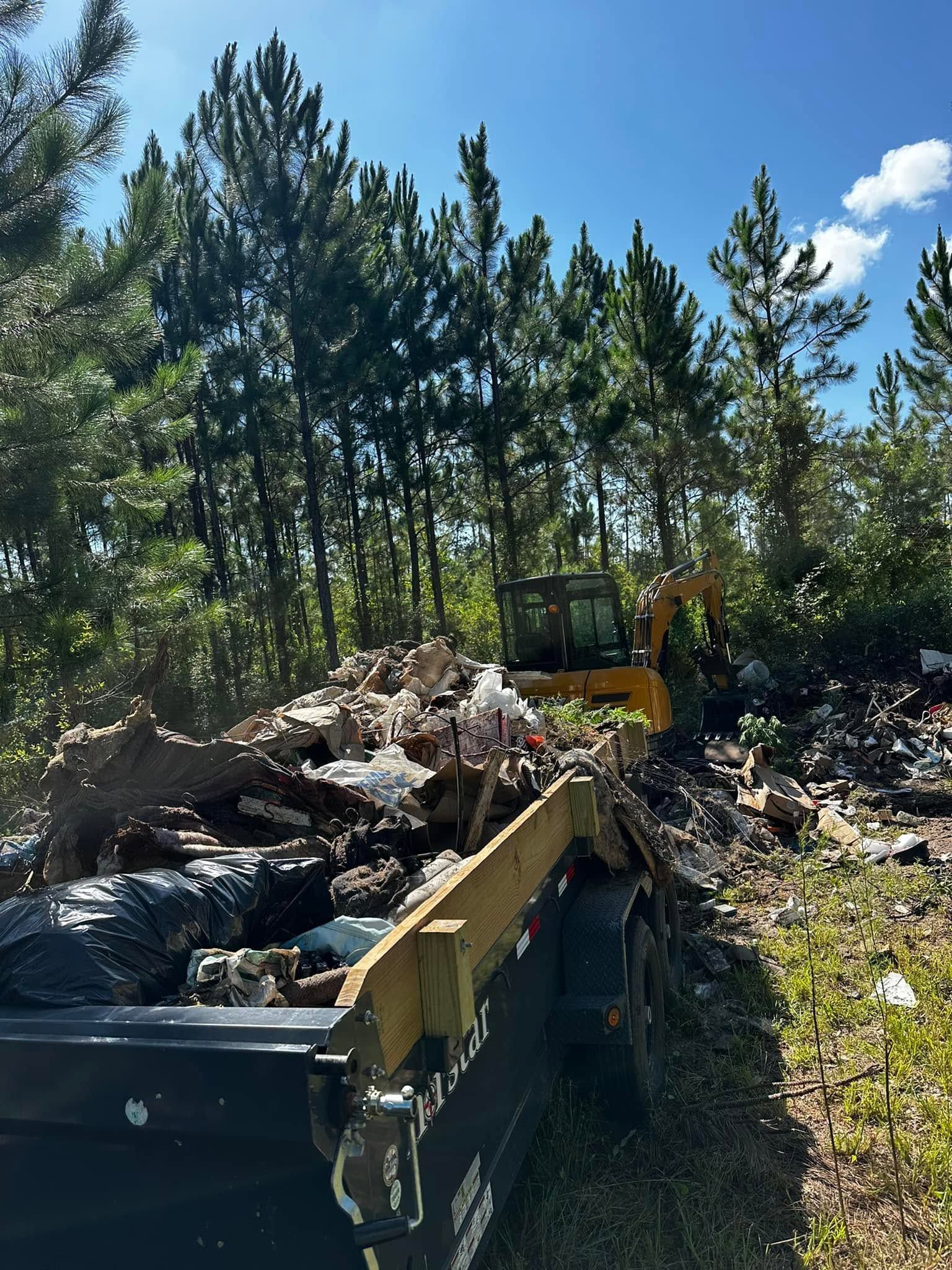 Construction debris piled in a dumpster beside a dirt road with pine trees under a blue sky