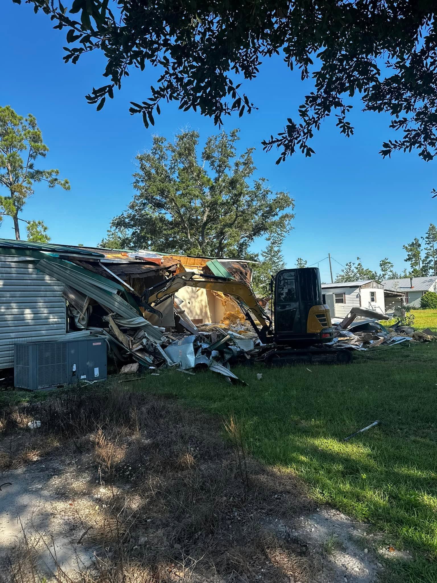 Collapsed mobile home in a yard with debris, a dumpster, and trees under a blue sky