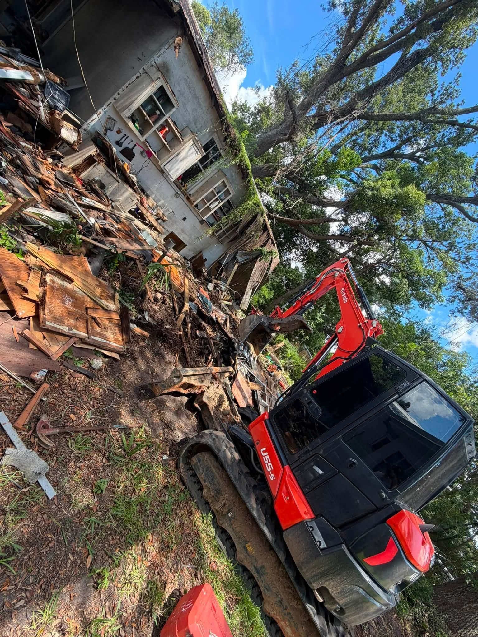 Backhoe clears debris beside a damaged house under blue sky.