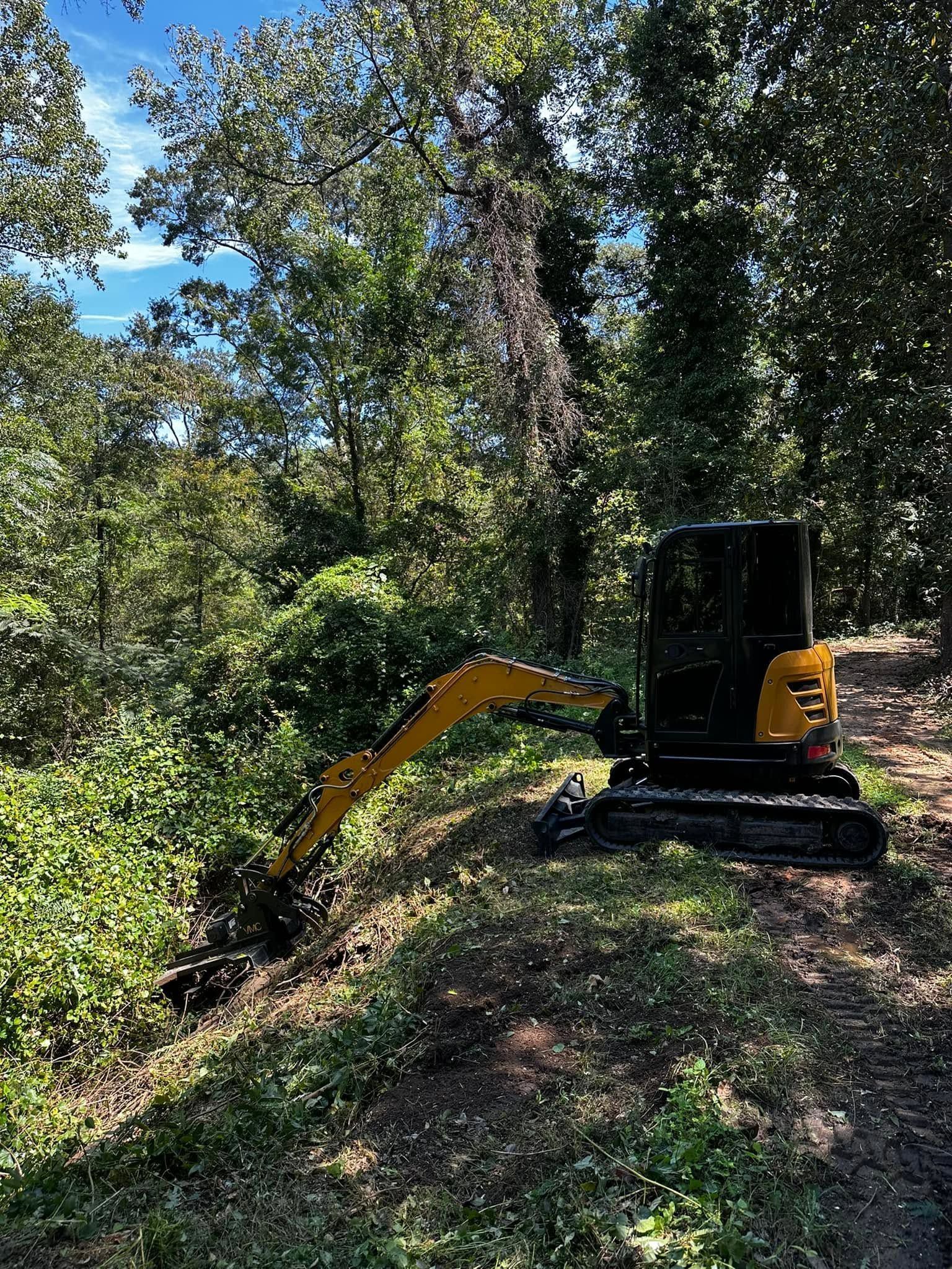 Yellow mini excavator clearing brush in a wooded area under a sunny sky
