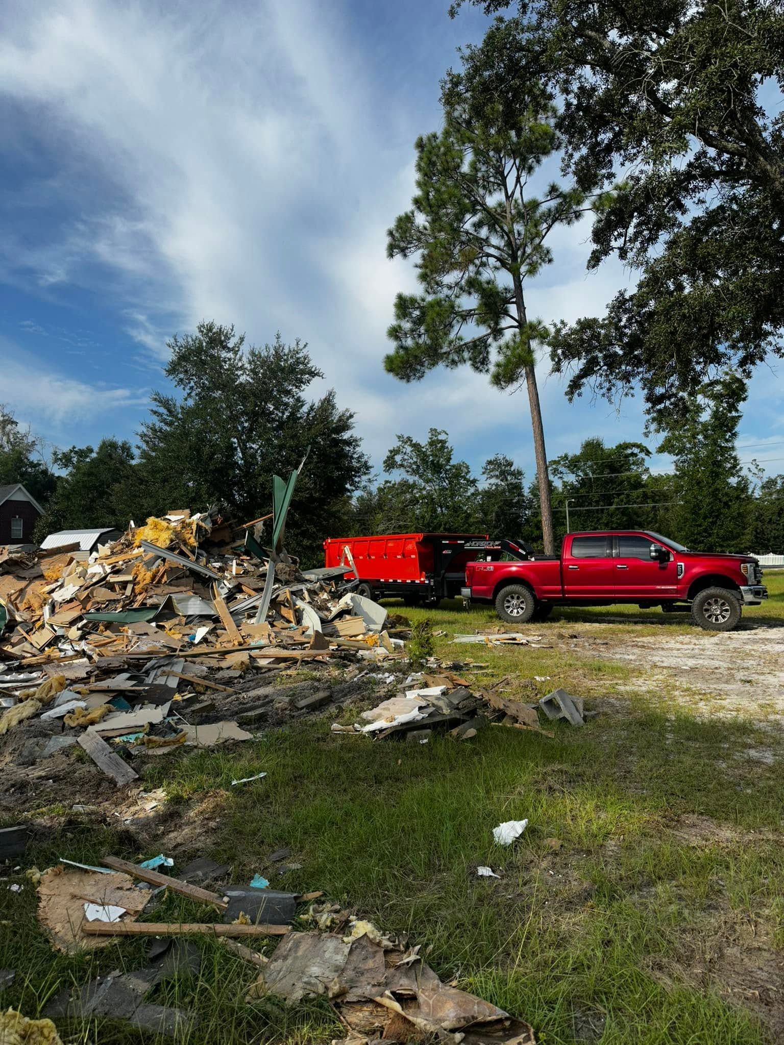 Red pickup and trailer beside a large pile of debris under a cloudy sky, with trees in the background