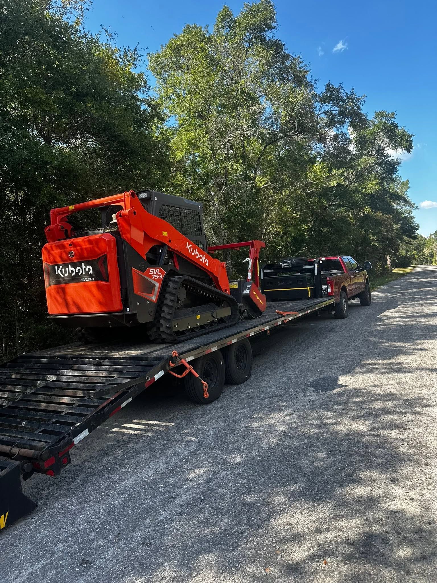 Orange skid-steer loader on a flatbed trailer beside a gravel road under trees