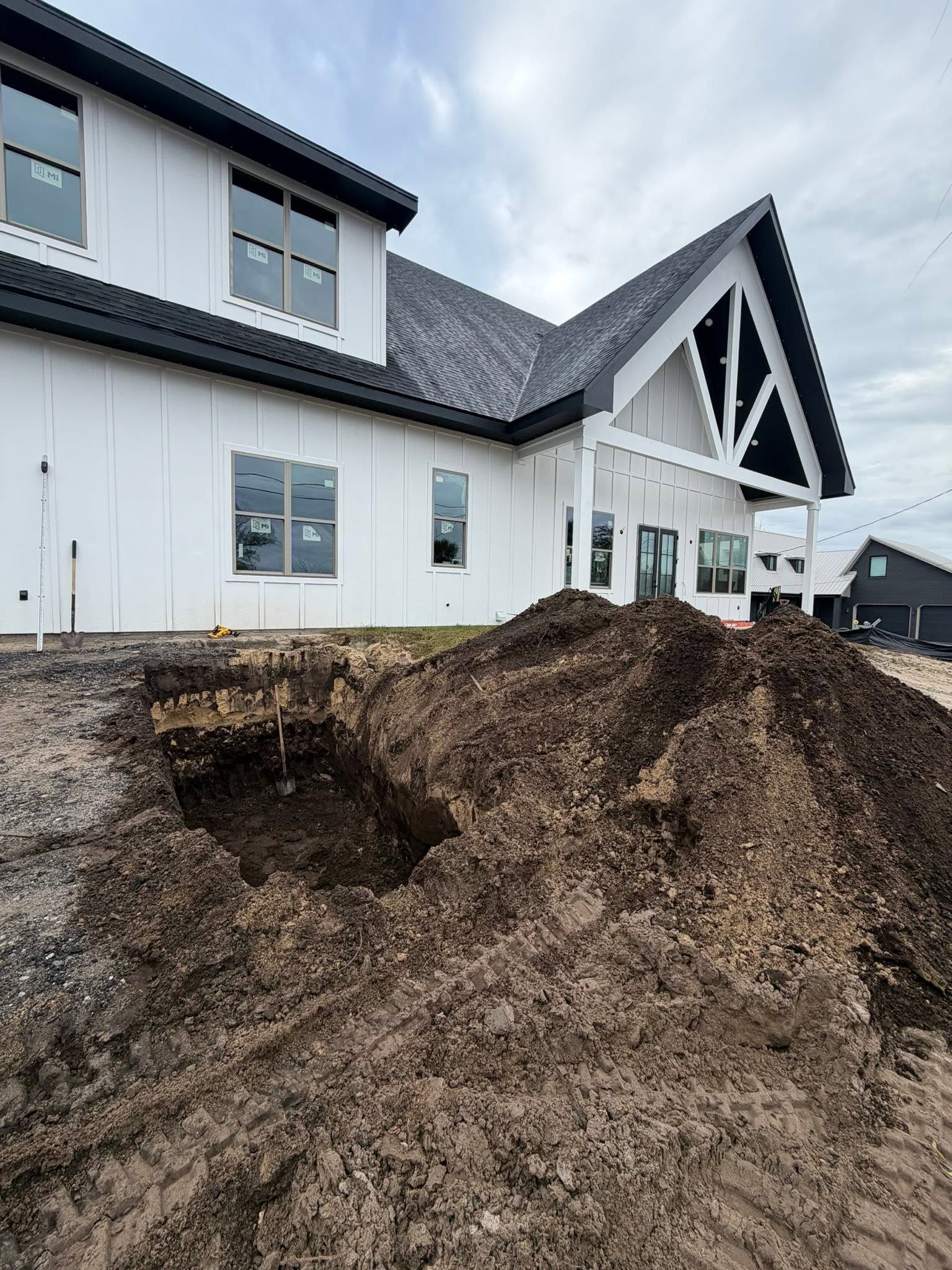 Large dirt mound and excavation hole in front of a white house under cloudy skies