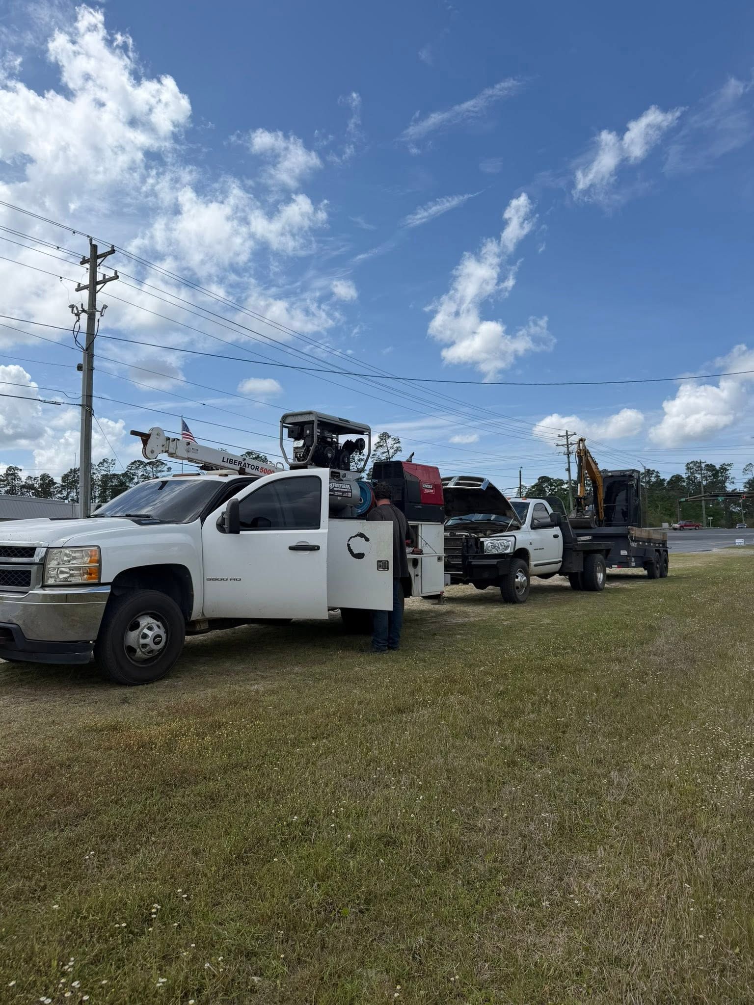 White work truck and trailers parked on grass under a blue sky with scattered clouds.