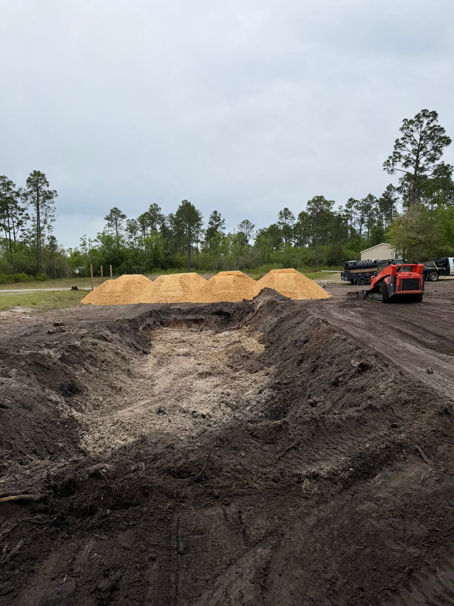 Large soil and gravel piles at a construction site with a red skid steer on the right.