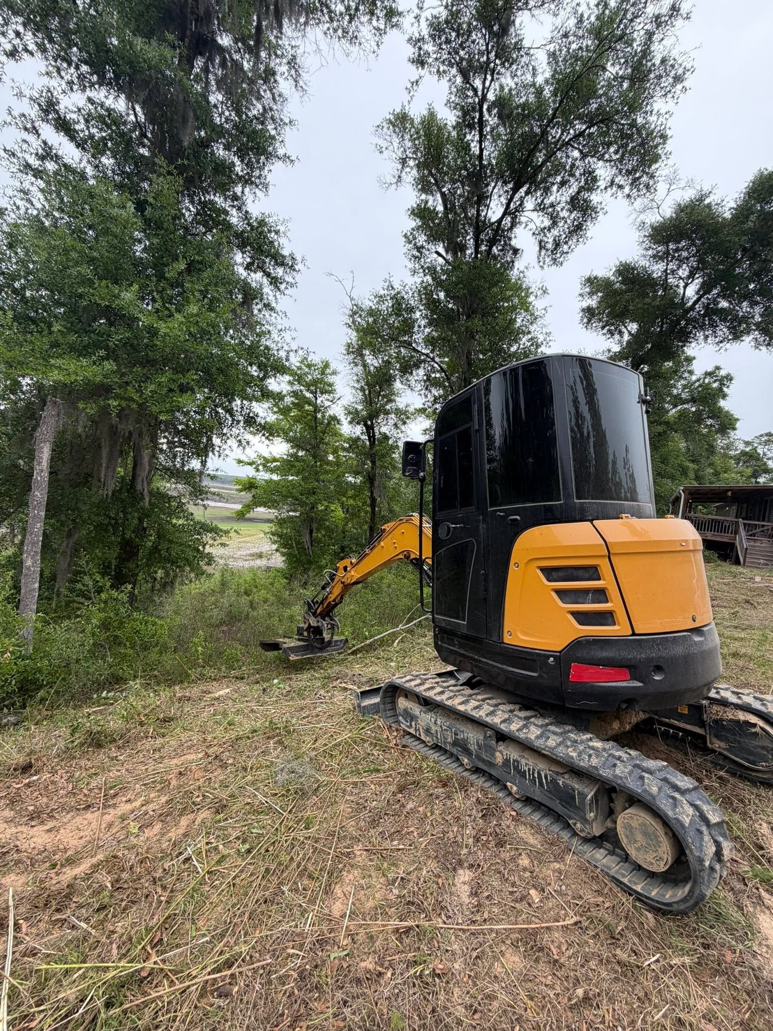 Yellow mini excavator on tracks clearing brush in a wooded area.