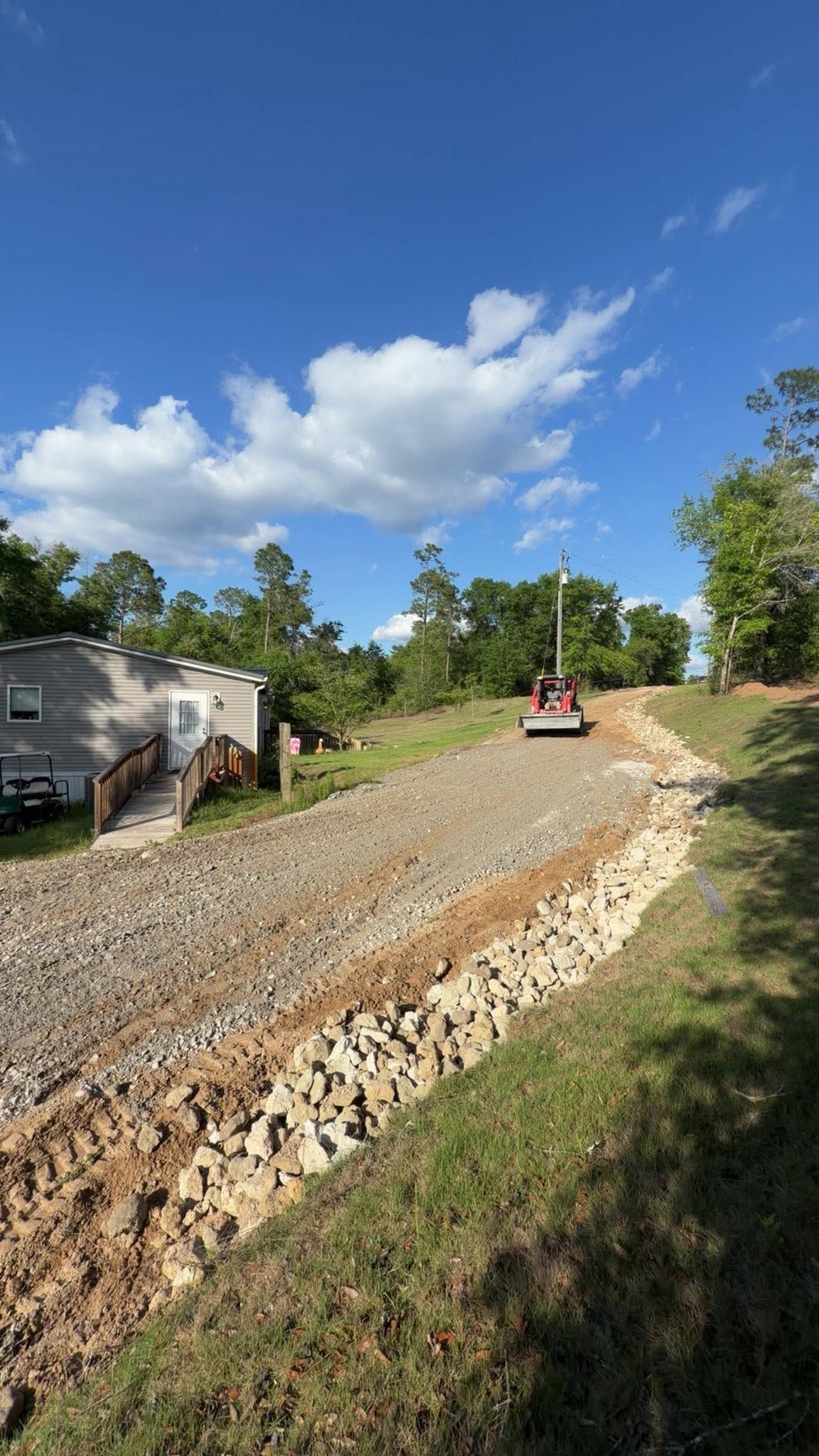 Gravel driveway under construction with an orange tractor, beside a house and trees under a blue sky.