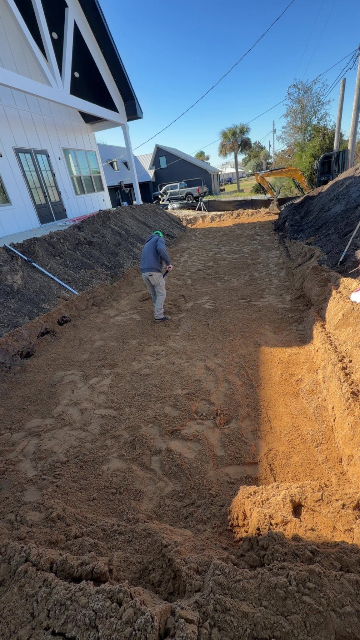 Worker in a muddy trench beside a house under construction, with dirt piles on both sides.