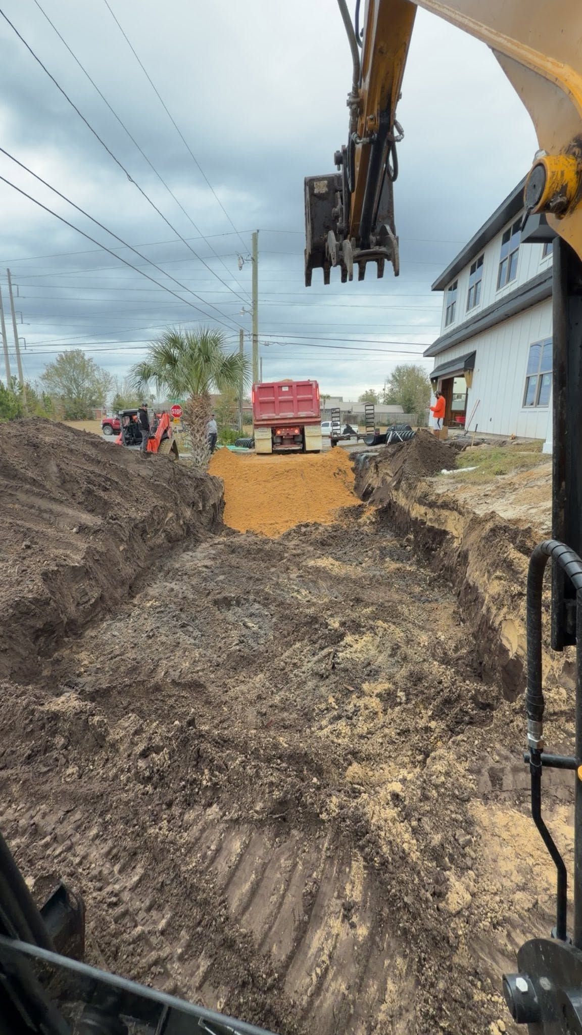 Excavator digging a trench beside a house on a cloudy day, with dirt piles and utility poles nearby