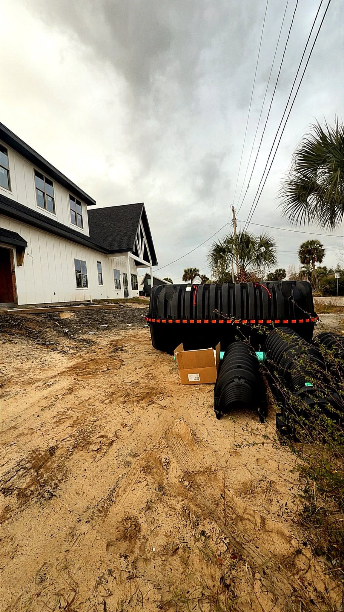Rural road construction with muddy gravel, a black fence, white houses, and overhead power lines under cloudy sky