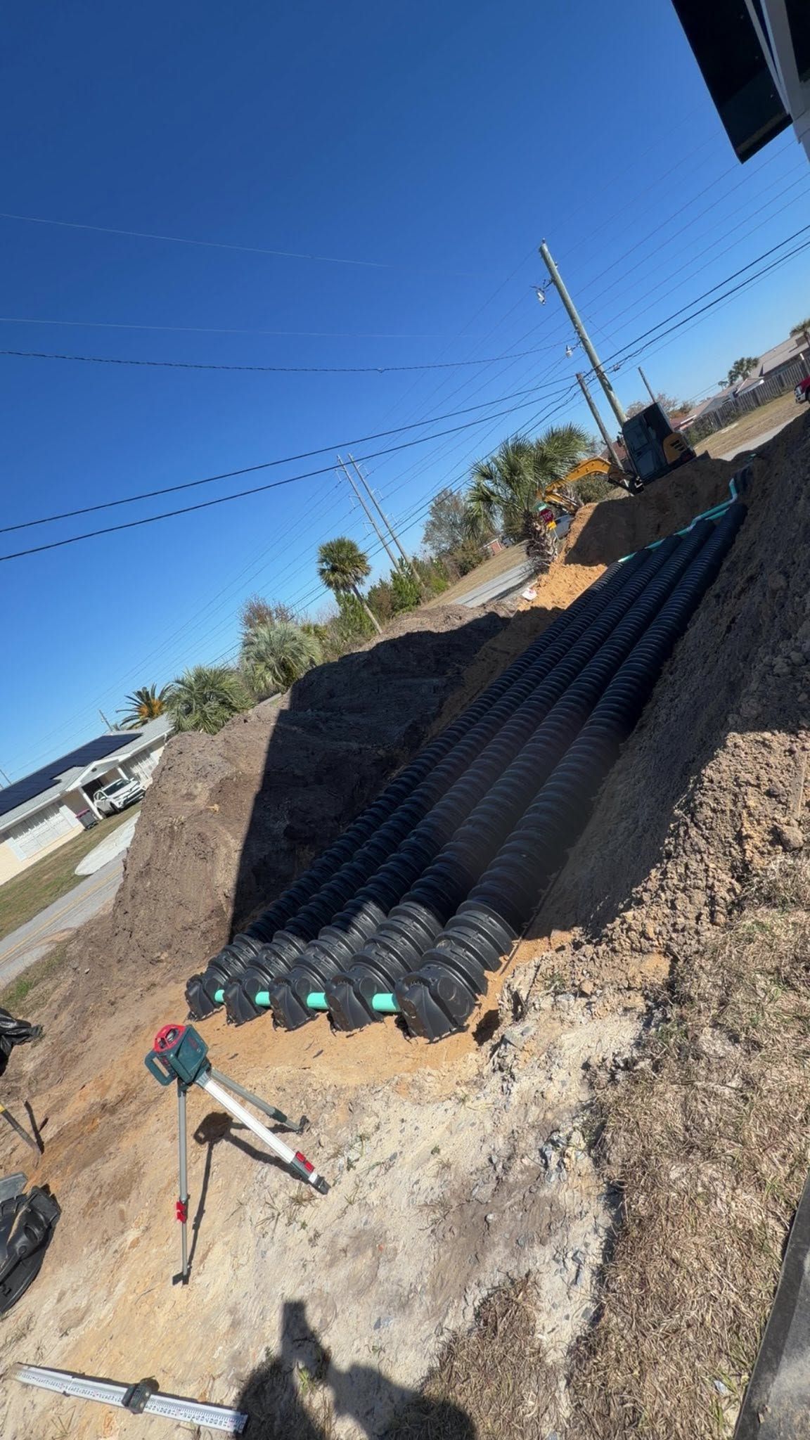 Plastic drainage pipes installed along a gravel slope beside a dirt road under a clear blue sky