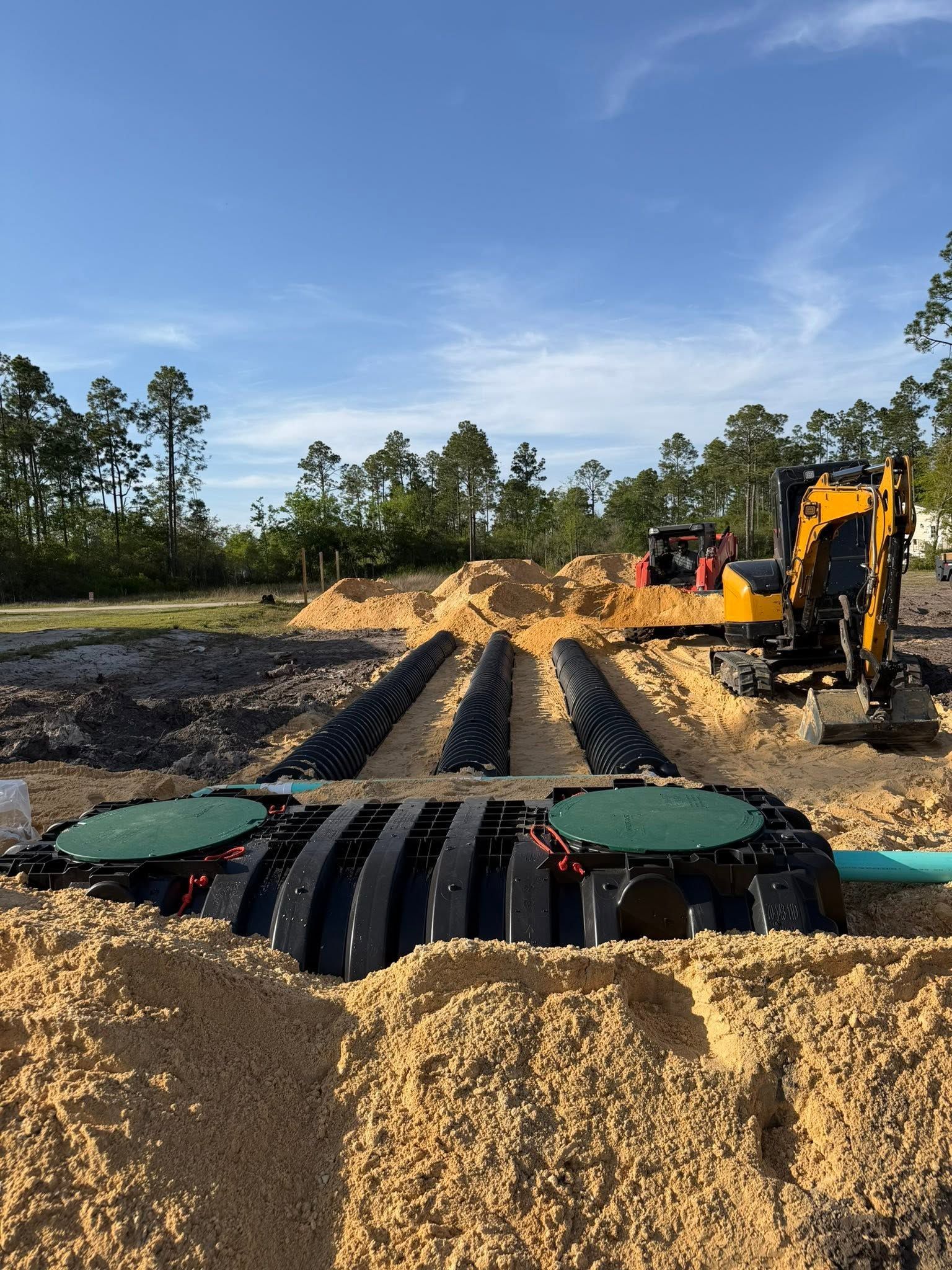 Septic tank installation site with yellow excavator and exposed black pipes in a sandy clearing