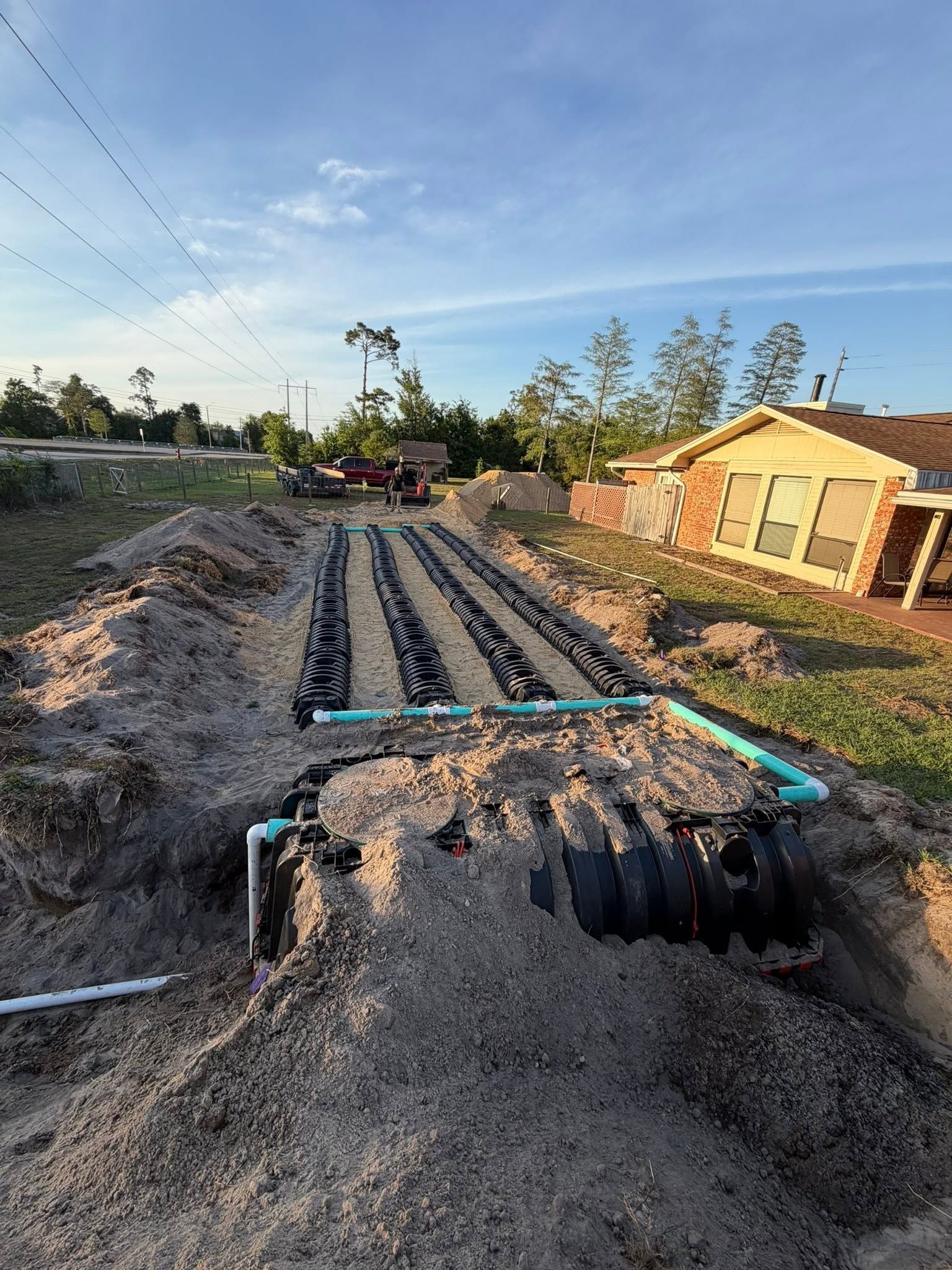 Trenches and blue pipes laid across a gravel yard beside a house at sunset