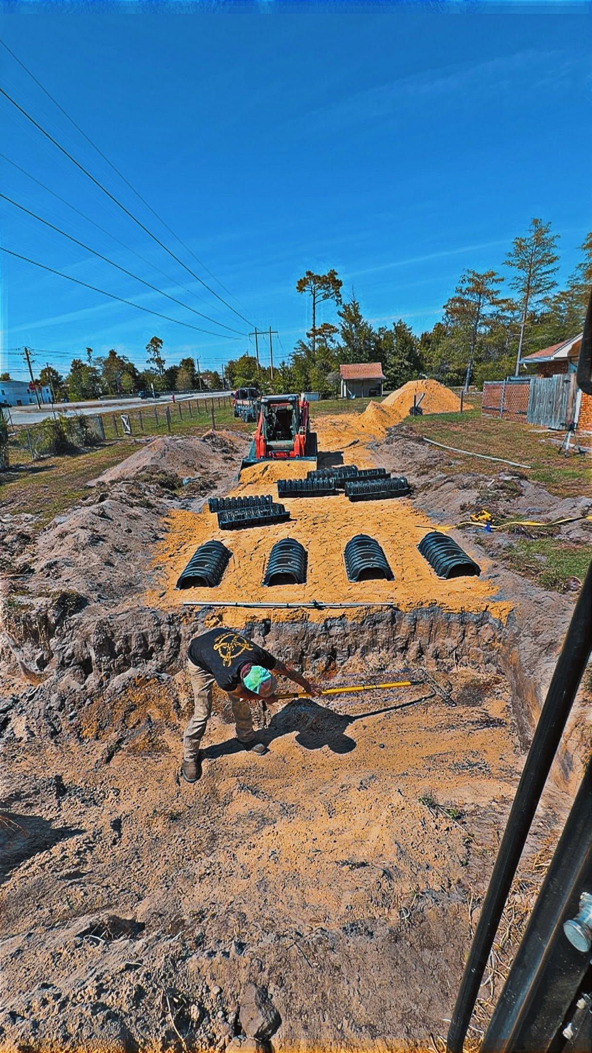 Yellow tarp with black bags on a road, with a small excavator and dirt piles under a blue sky.
