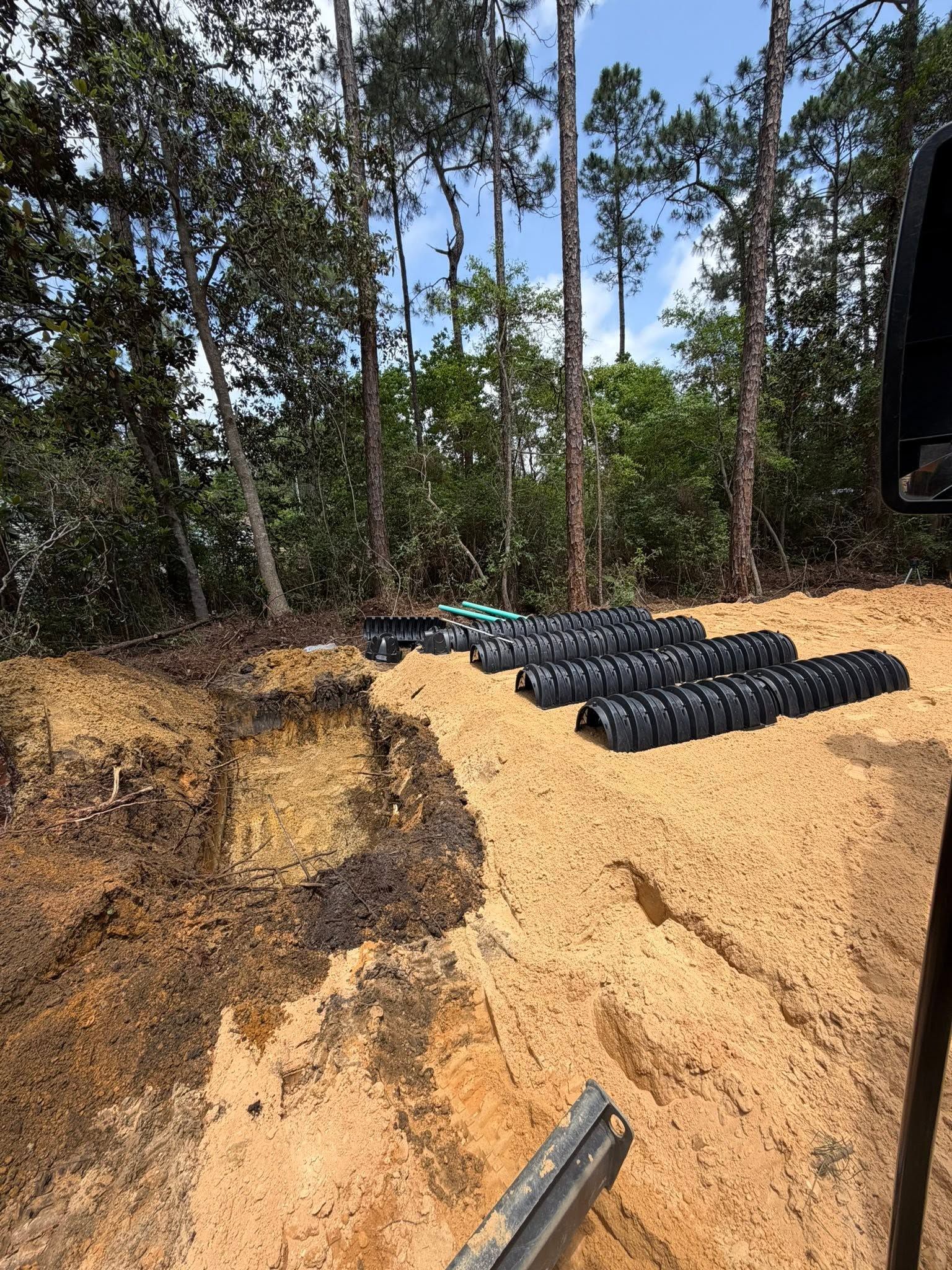 Excavated dirt site with rows of black pipes on a sandy slope beside trees.