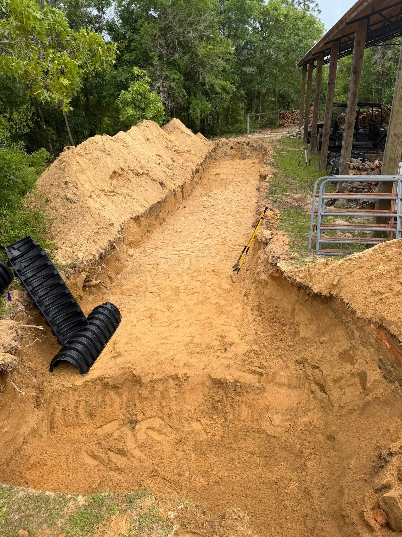 Large pile of sand beside a black drainage pipe at a muddy construction site with trees and a shed.