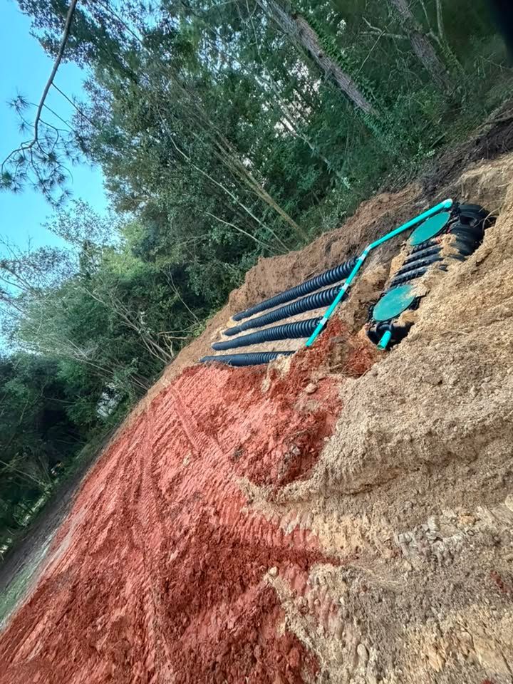 Terraced hillside excavation with red soil, black pipes, and a turquoise hose beside a wooded area