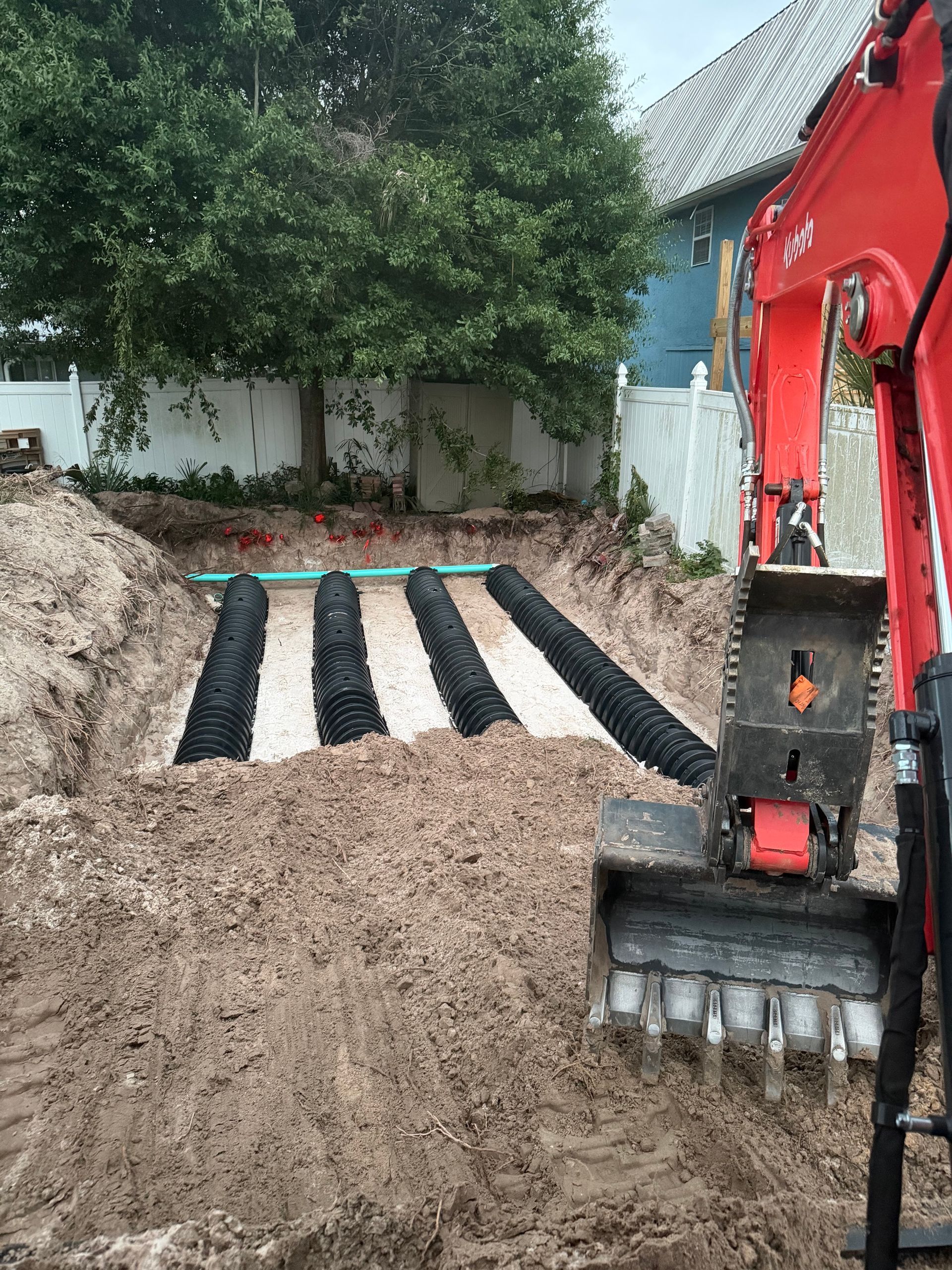 Excavator digging a trench beside rows of black drainage pipes in a backyard construction site