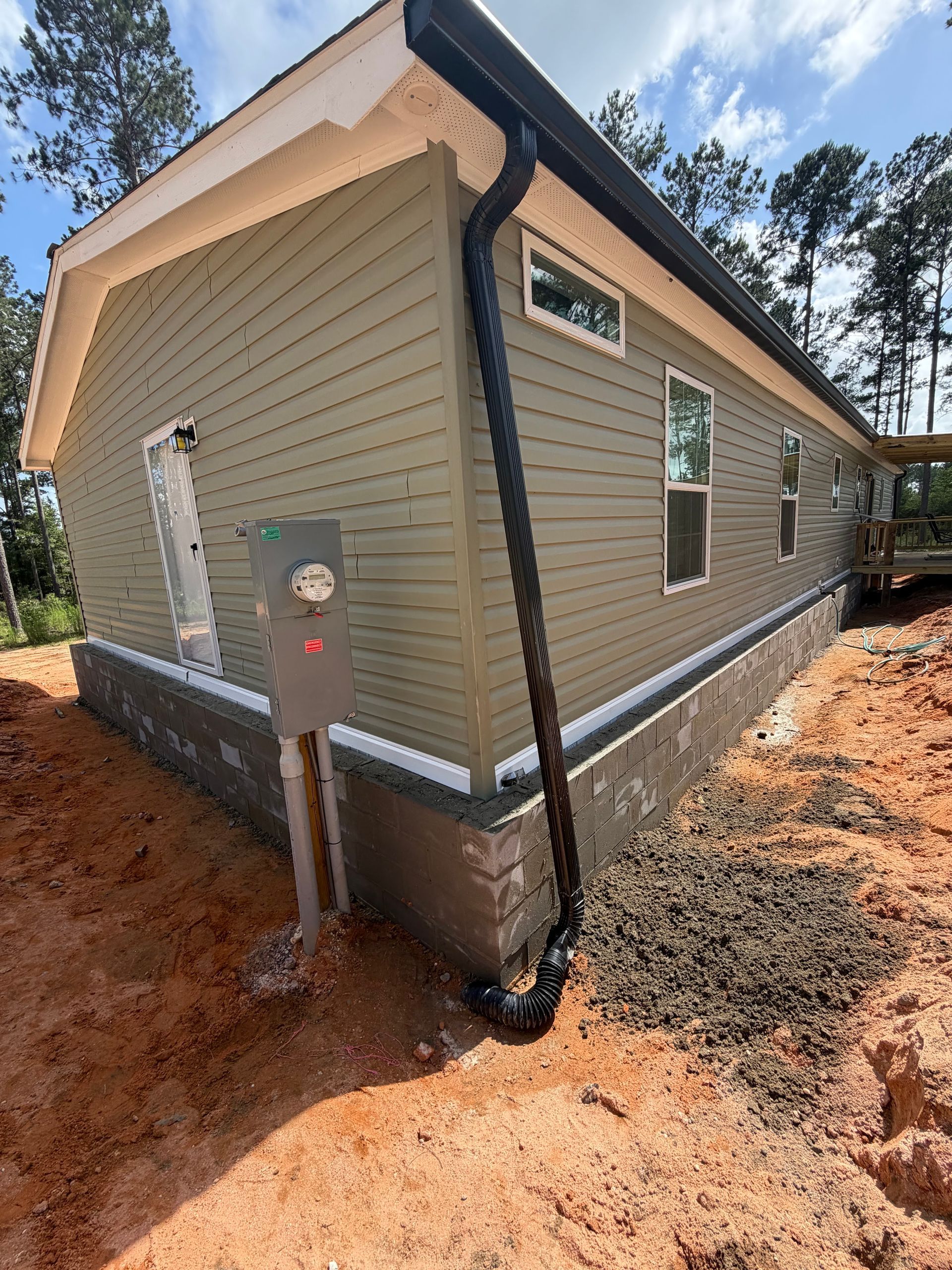 Side view of a beige house with white trim, black gutter, and utility meter on a dirt lot under a blue sky.