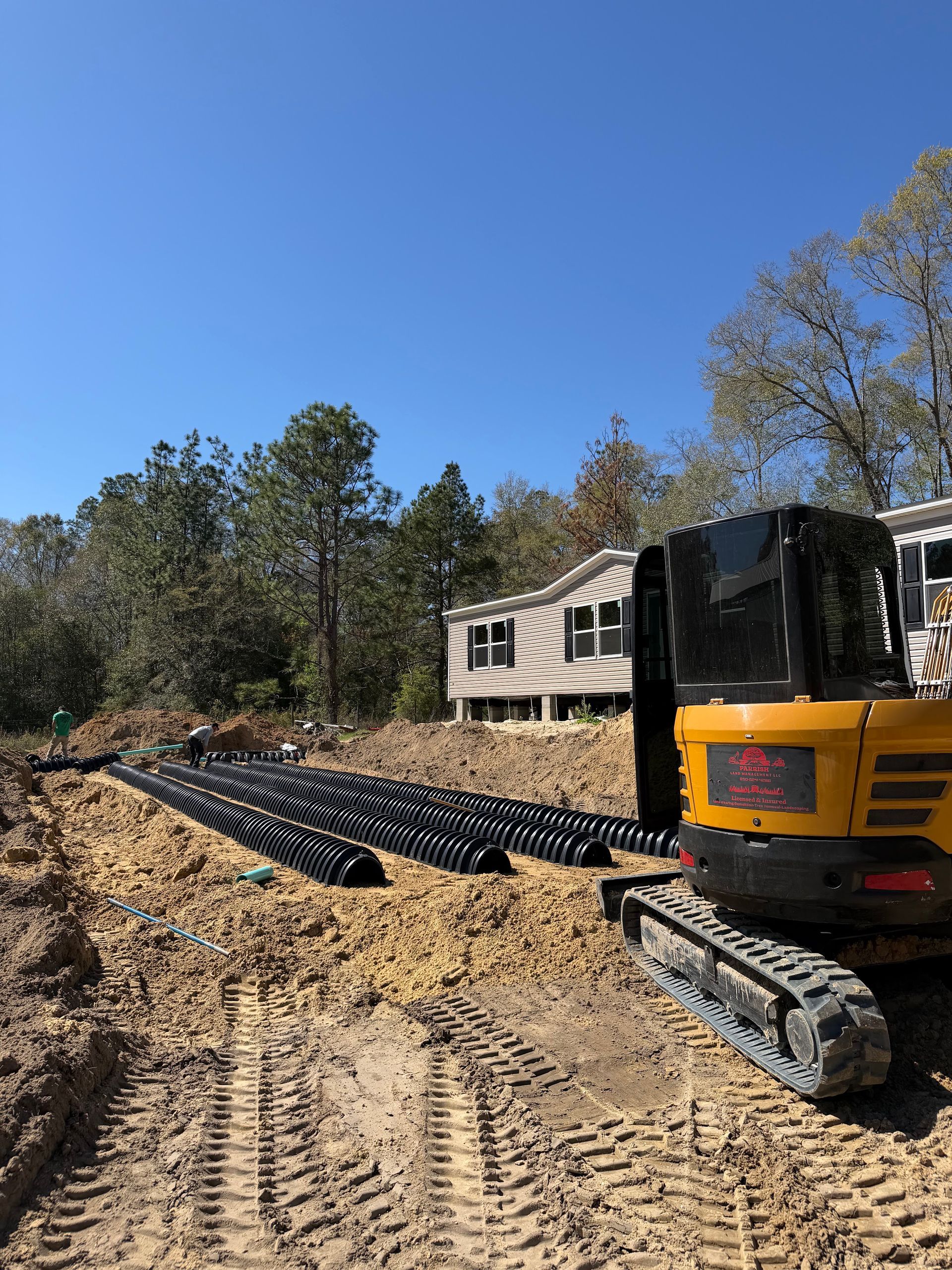 Yellow excavator laying black pipe beside a mobile home on a dirt construction site under a blue sky