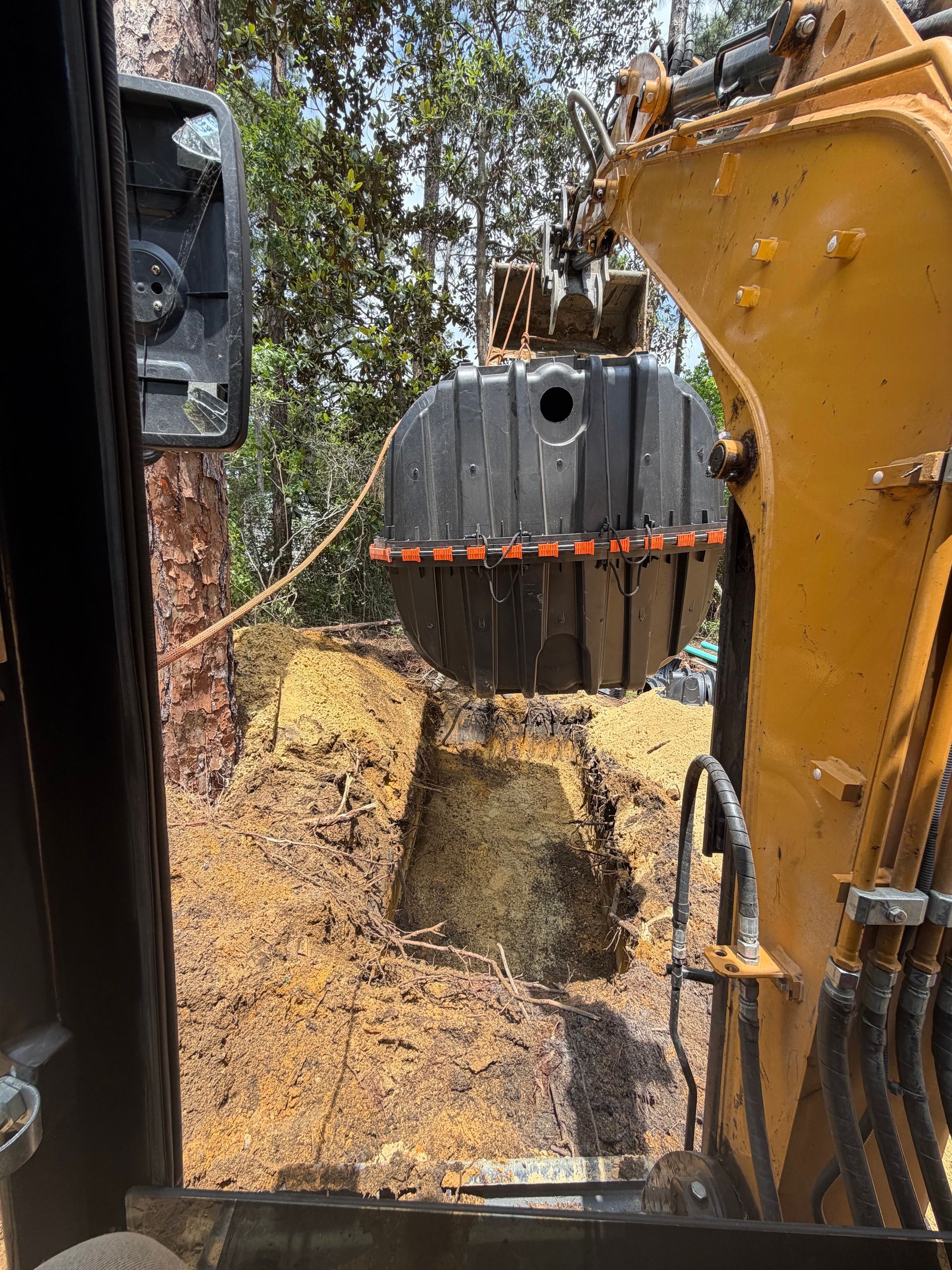 Excavator bucket lowering soil beside a black tank in a wooded construction site