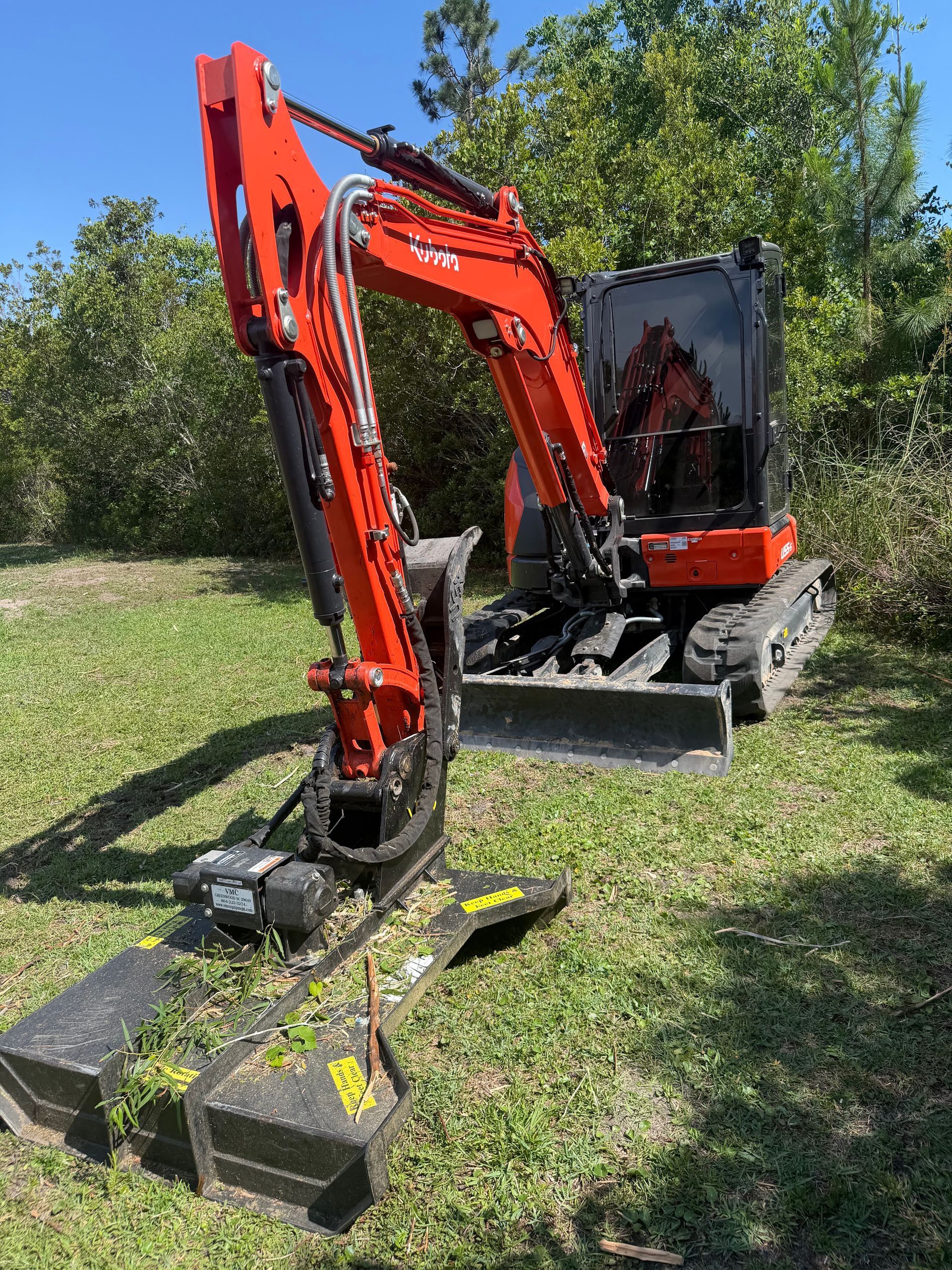Orange mini excavator with brush mower attachment on grassy ground outdoors.