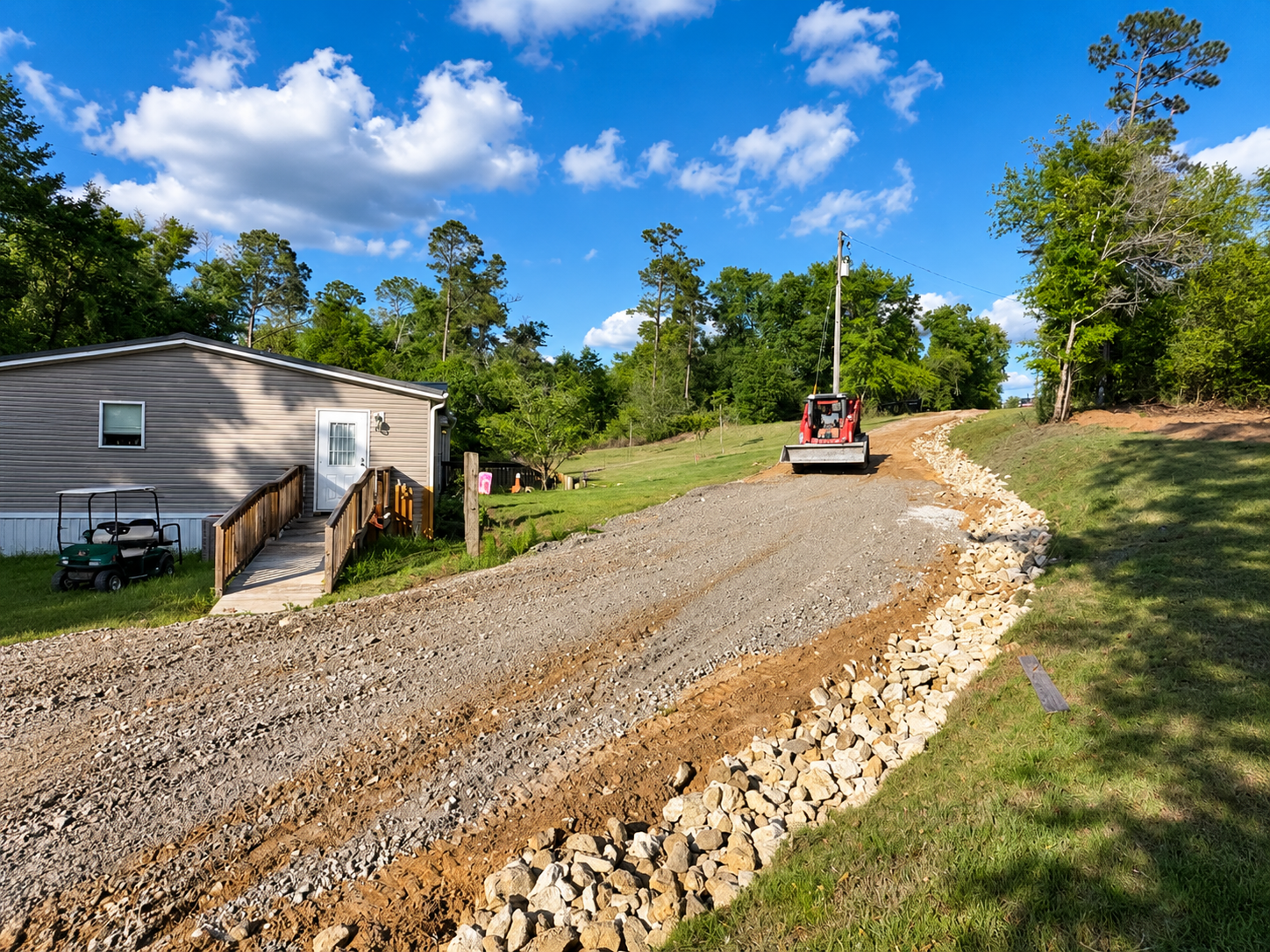 Gravel driveway under construction beside a house, with a small red excavator and rock edging on a sunny day
