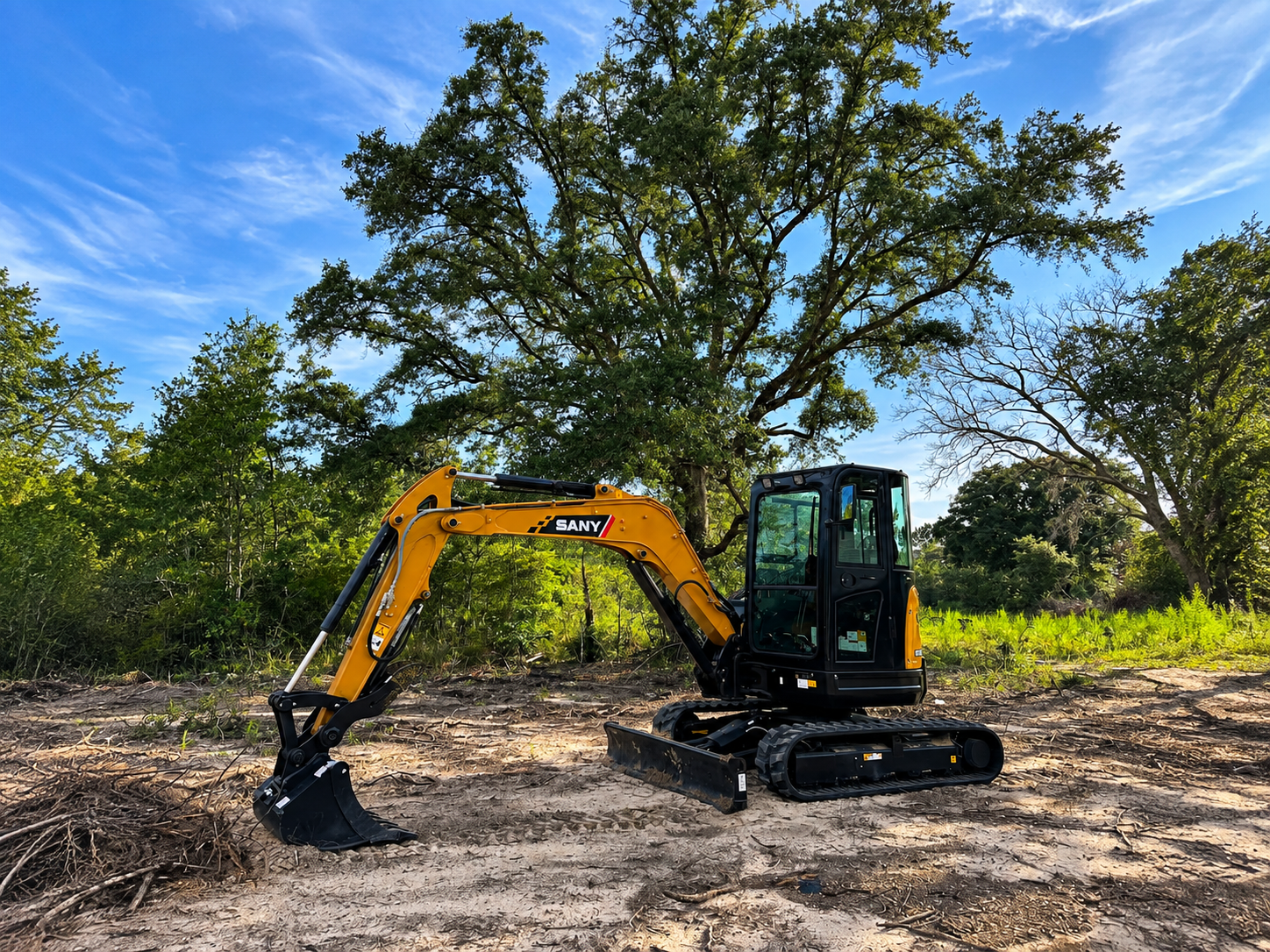 Yellow excavator clearing brush on a dirt lot under trees and a blue sky.