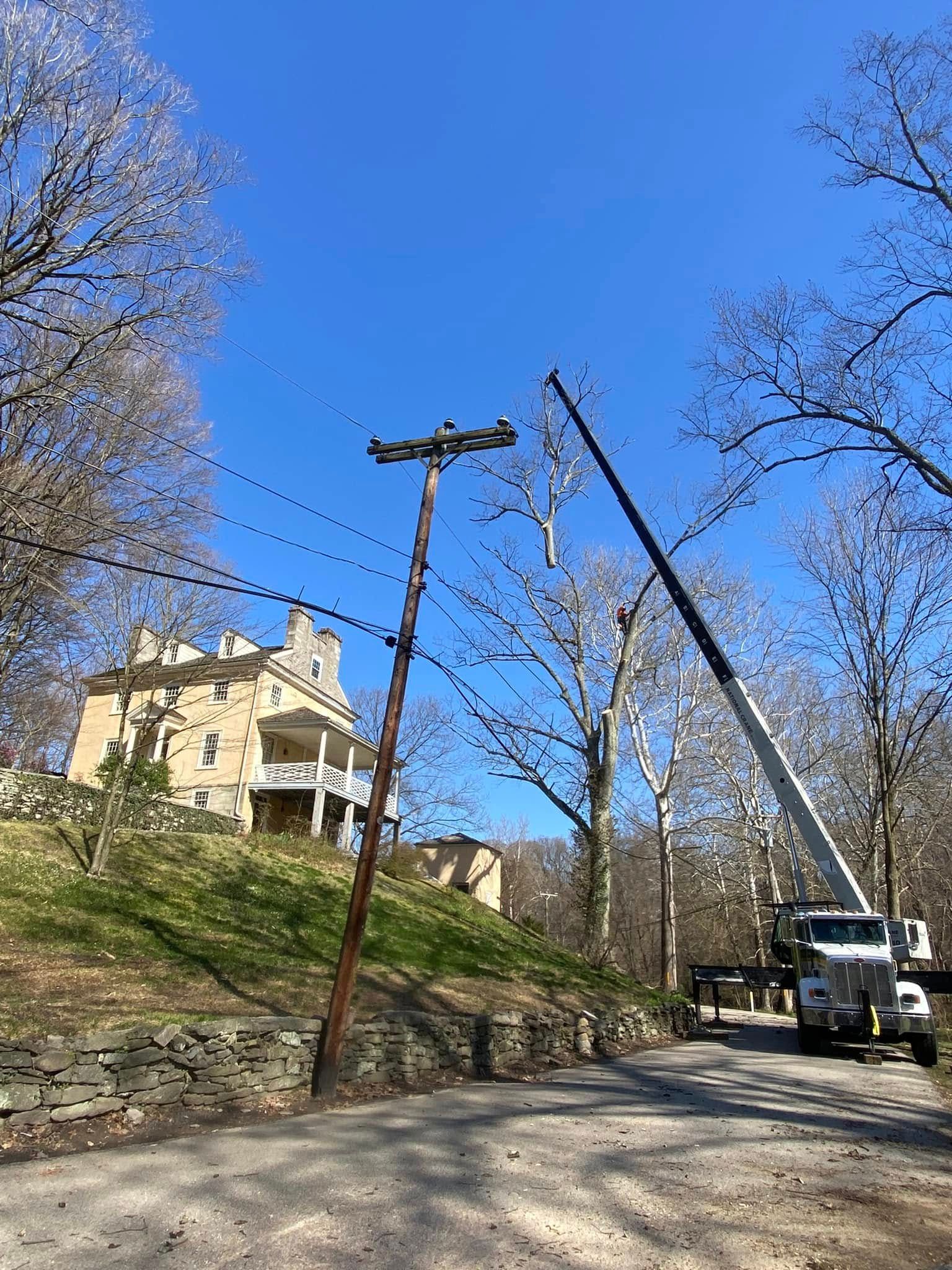 Utility trucks and power poles repairing overhead lines on a tree-lined road beside a yellow house