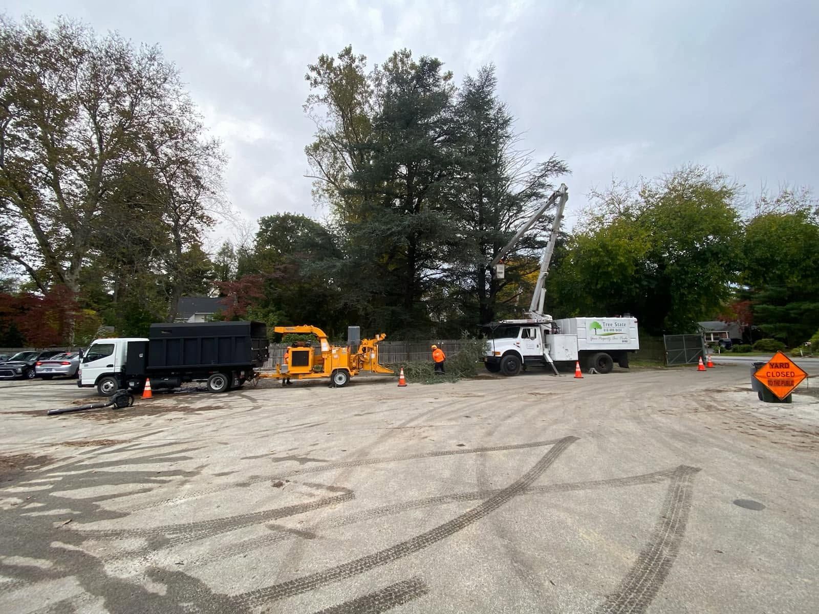 Roadwork staging area with trucks, orange equipment, and workers in a large paved lot.