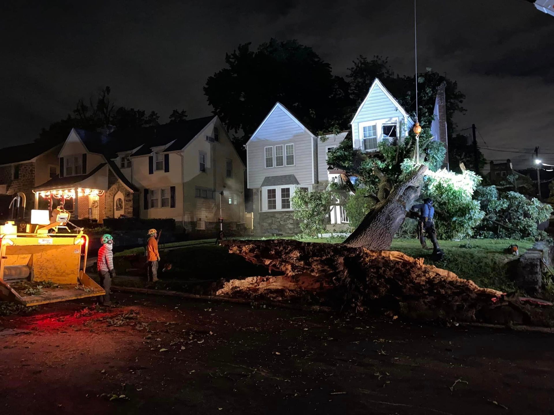 Nighttime storm damage with uprooted tree blocking a suburban street, workers and a utility vehicle nearby.