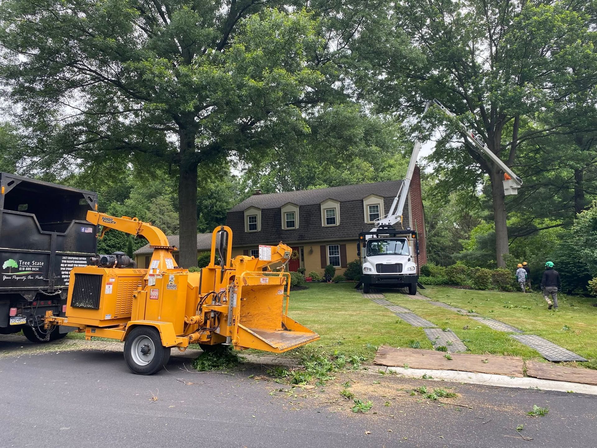 Orange wood chipper in a tree-lined yard beside a house, with a truck and white utility lift.