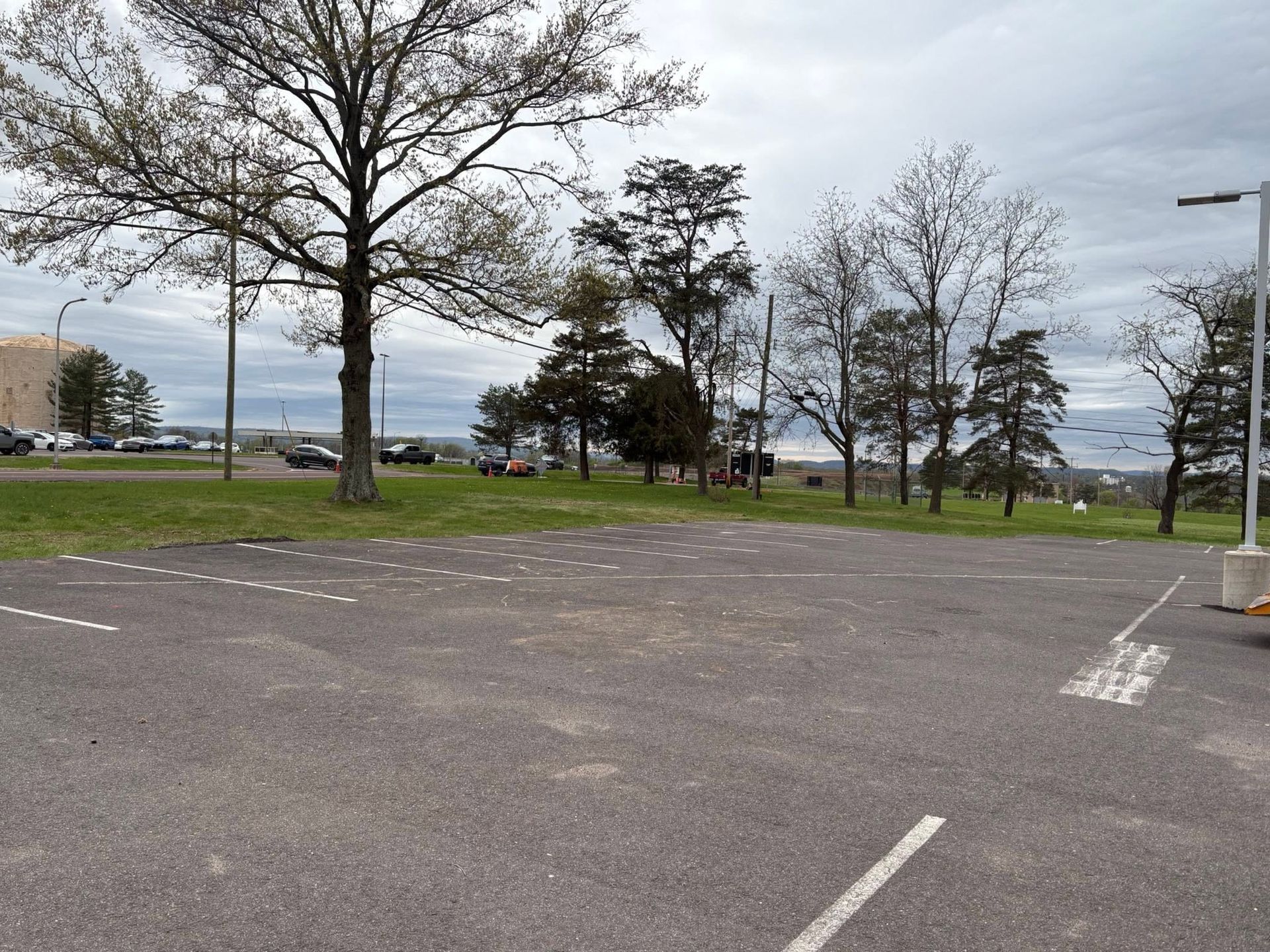 Empty parking lot by a grassy park under cloudy skies, with leafless trees and a distant water view.