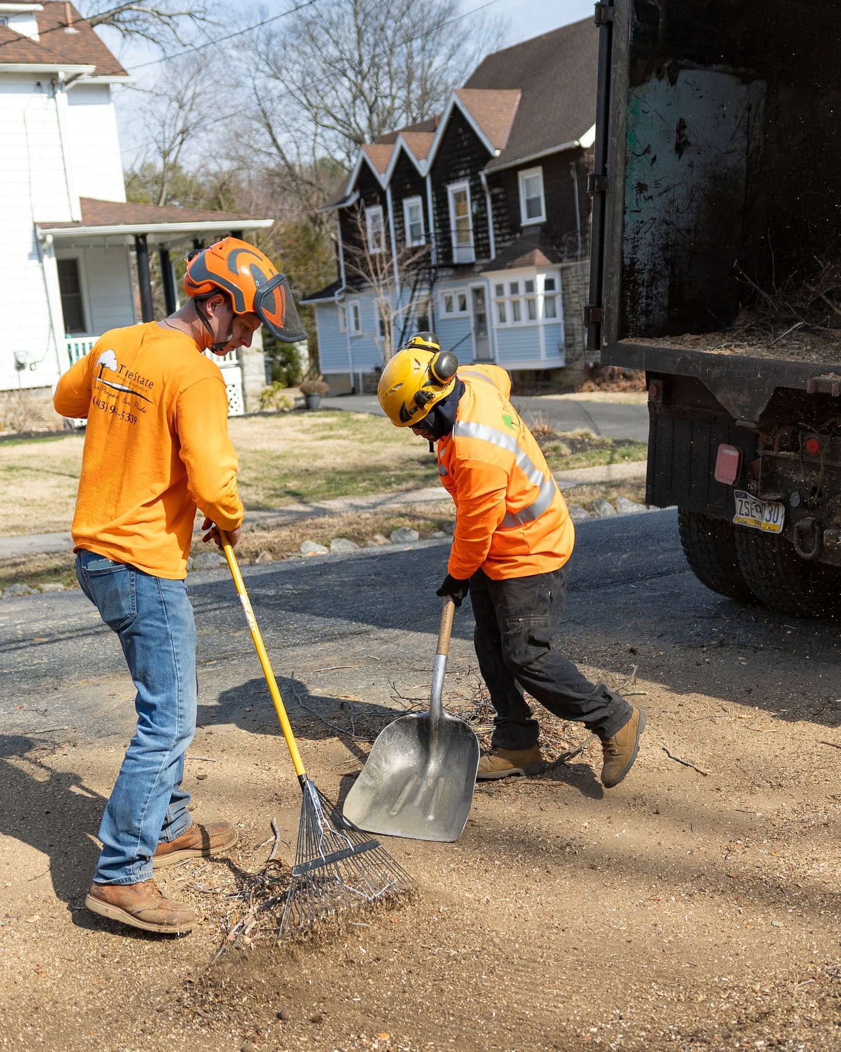 Two workers in orange safety gear shovel gravel beside a parked truck on a street.