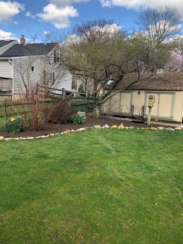 Backyard with green lawn, trees, flower bed, and two houses under a partly cloudy sky