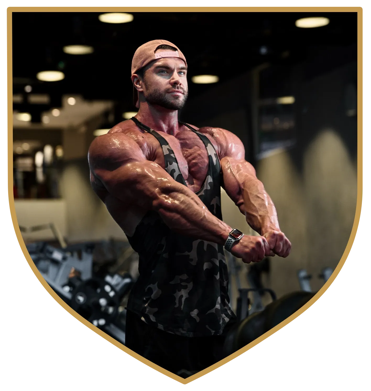 Muscular individual in a camouflage tank top and cap posing in a gym, highlighting arm and shoulder muscle definition.