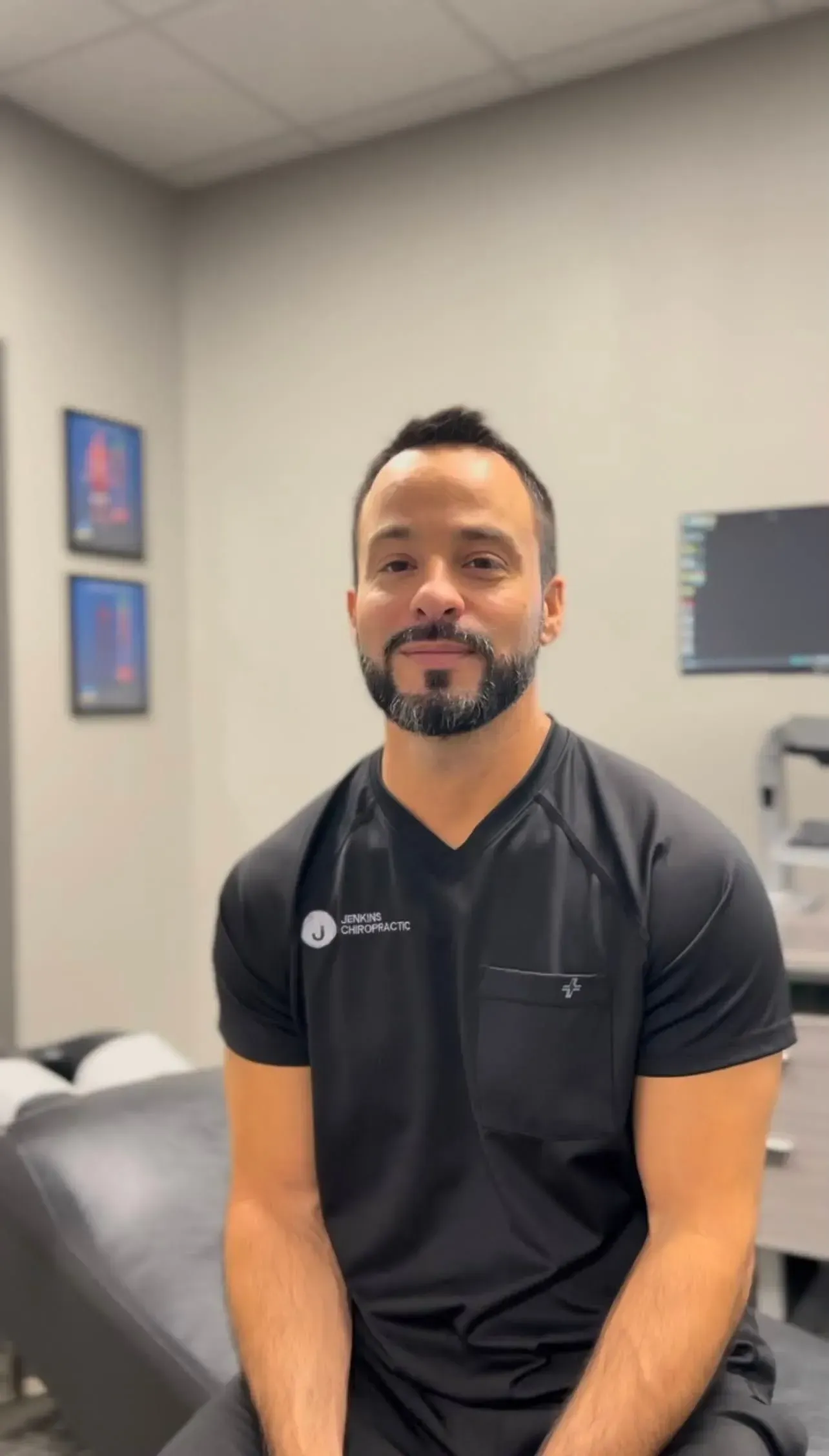 Man in black scrubs in office setting, smiling at camera.