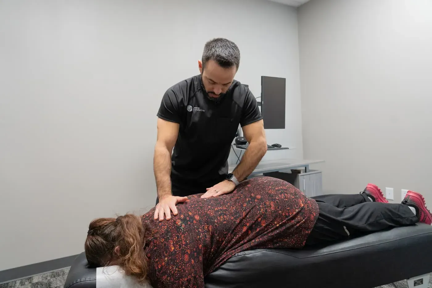 Chiropractor adjusting a patient's back on an examination table. Neutral-colored room.