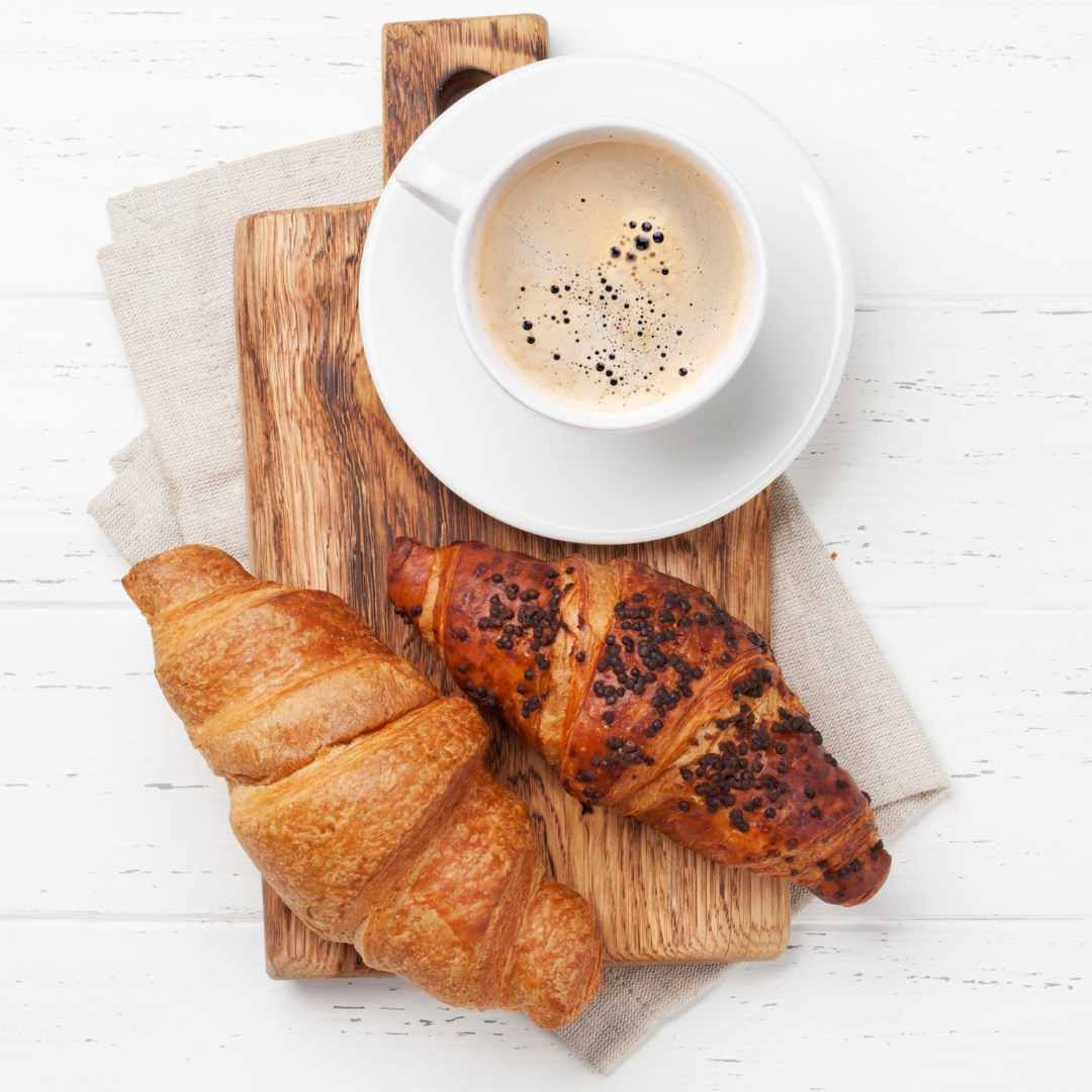 Croissants and coffee on a wooden board, atop a neutral-colored cloth, on a white wooden surface.