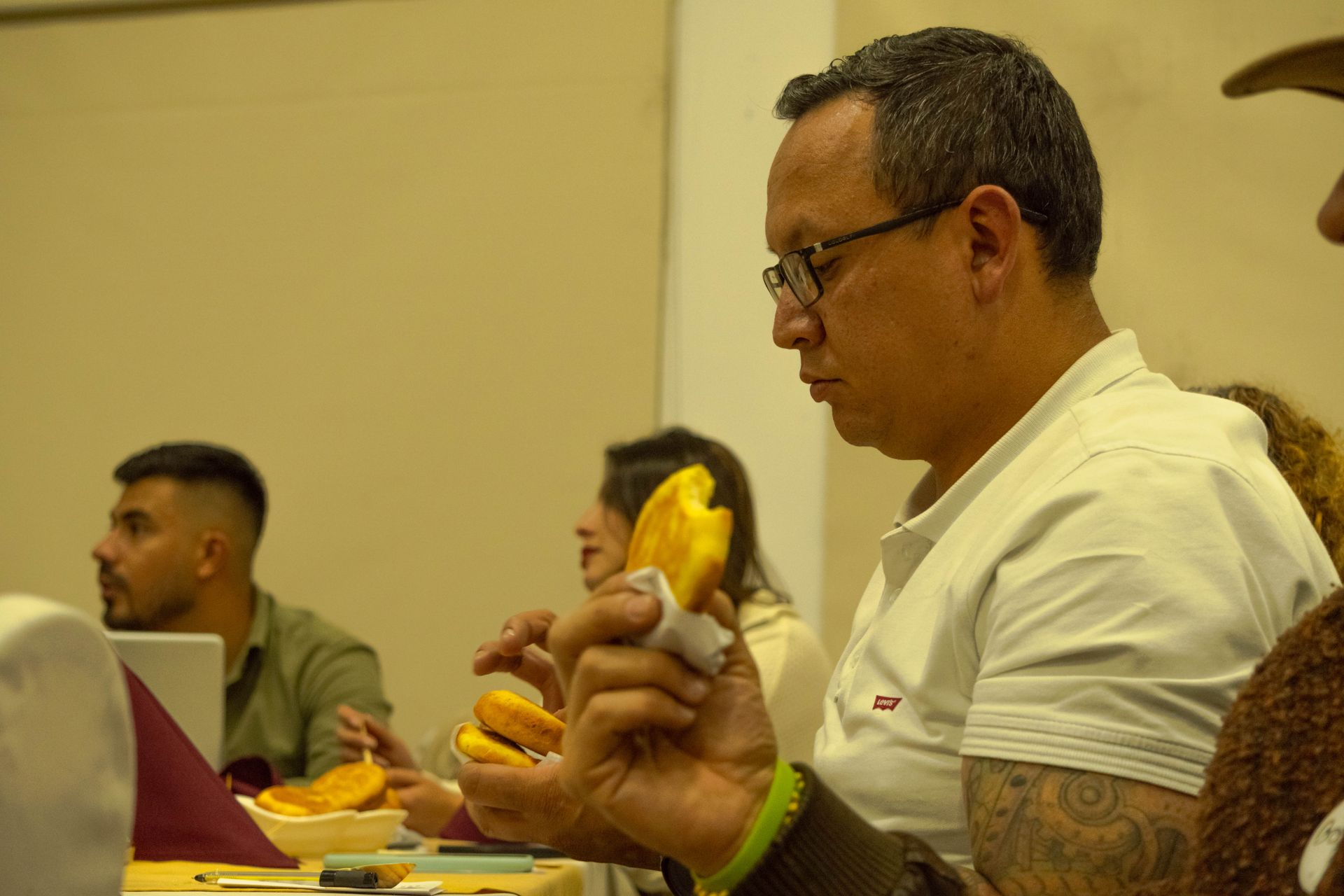 Man eating at a table, wearing glasses and a white shirt. Others are visible in a restaurant setting.