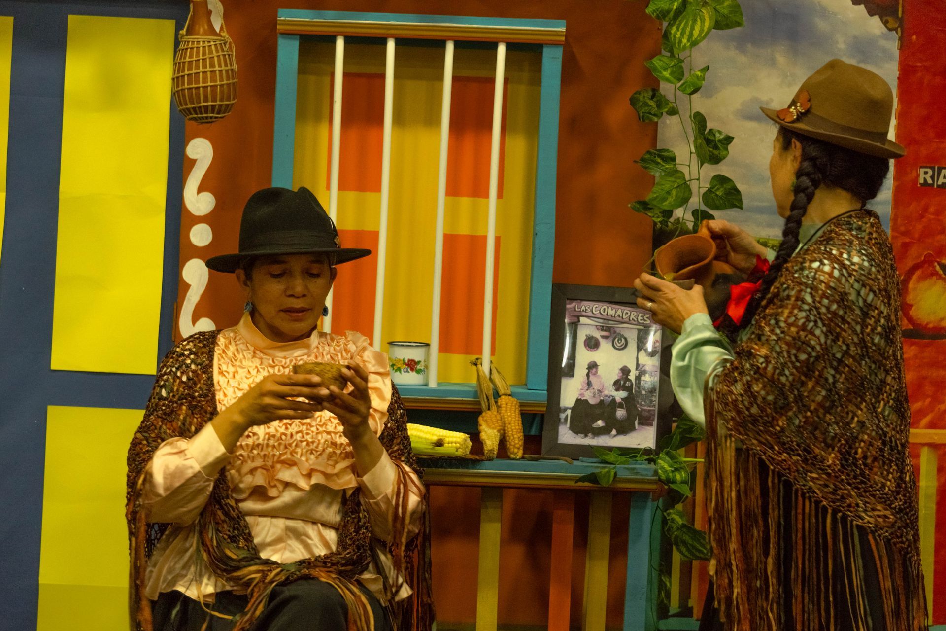 Two women in hats and shawls in a colorful room, holding cups. One is seated, the other standing.