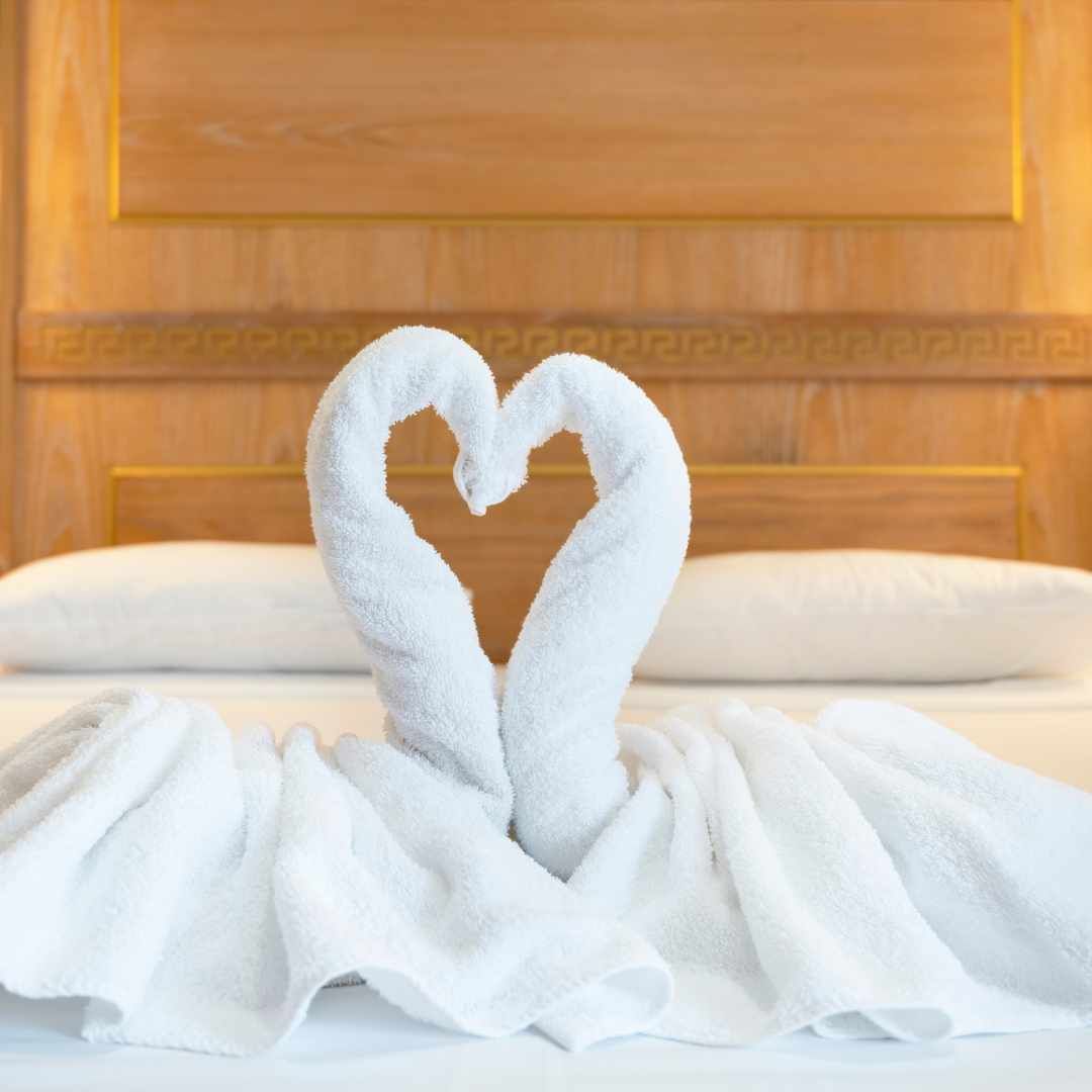 Two white towel swans in a heart shape on a made bed in a hotel room with a wooden headboard.