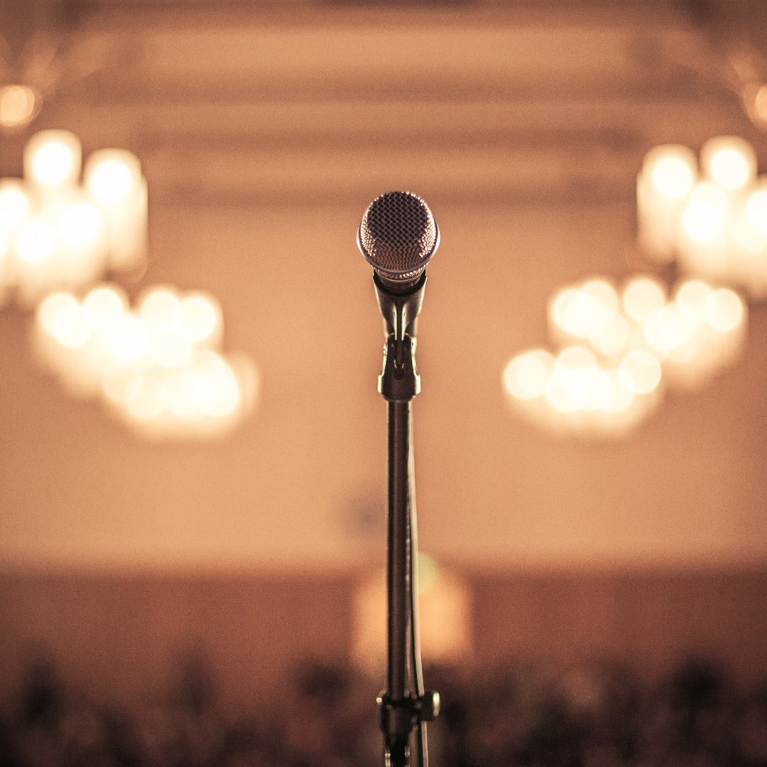 Microphone on a stand, centered in a warm-lit hall with blurred lights in the background.