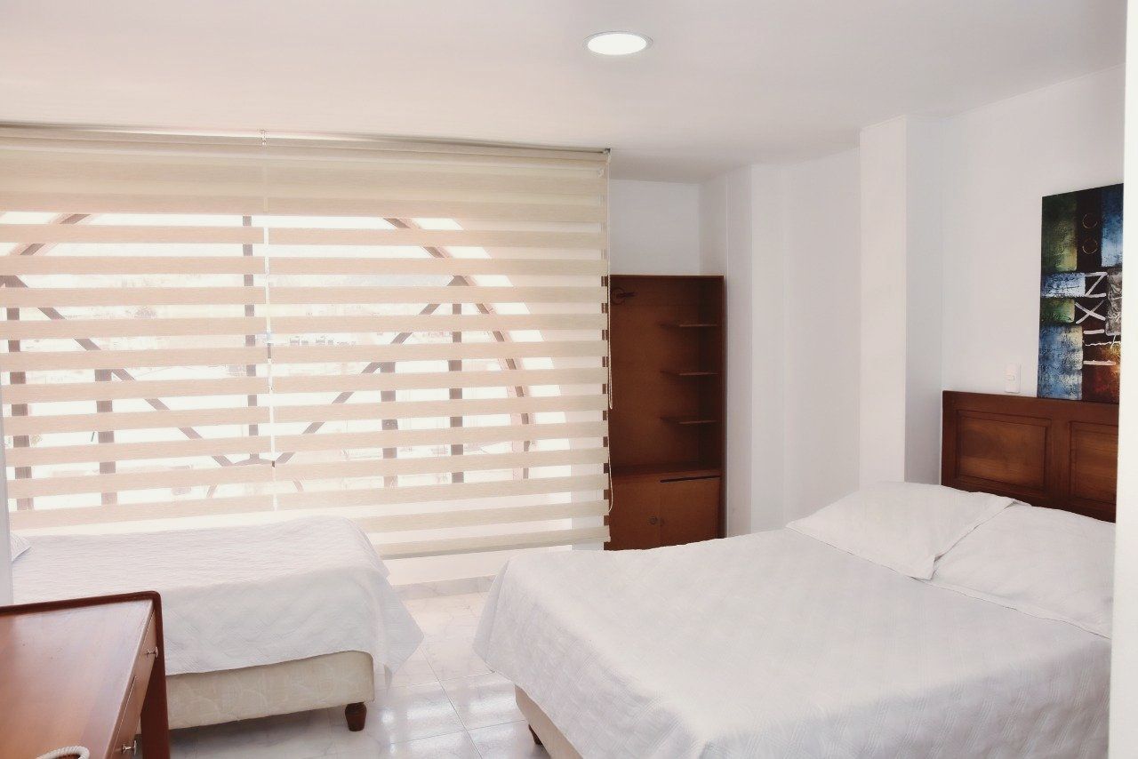 Bedroom with two beds, a dresser, and a tall wooden cabinet. White walls and window blinds.