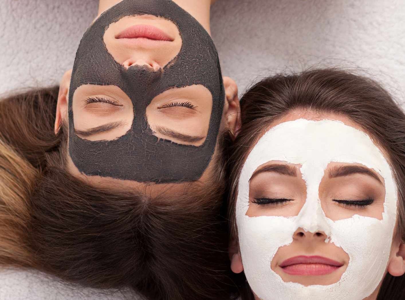 Two women with closed eyes, each with a facial mask: one black, one white. Lying side-by-side on white.