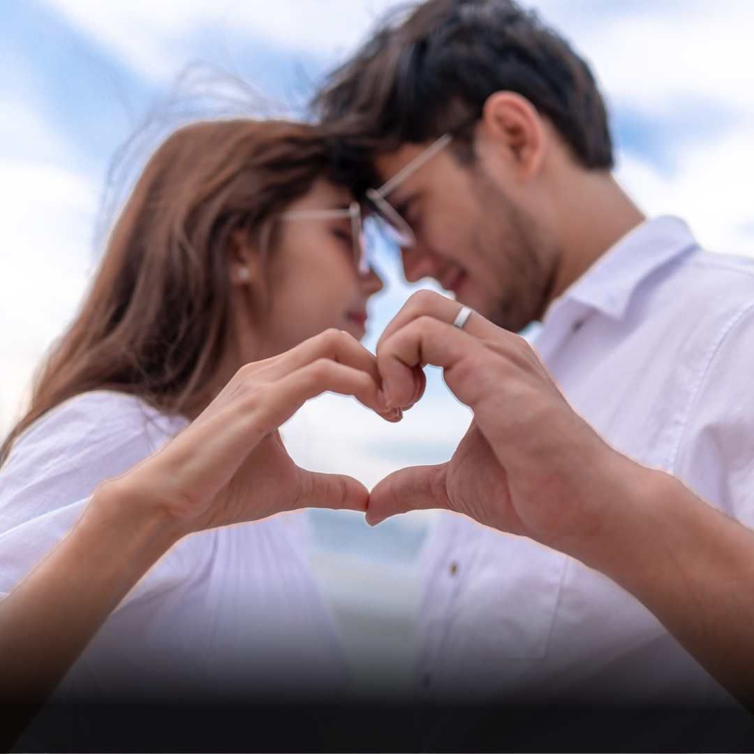 Couple forming a heart shape with their hands, foreheads touching, outdoors.