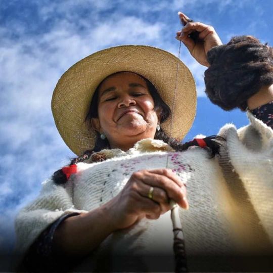 Woman in straw hat, knitting with yarn outdoors against a blue sky.