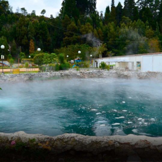 Steaming turquoise hot spring pool with a white building and green trees in the background.