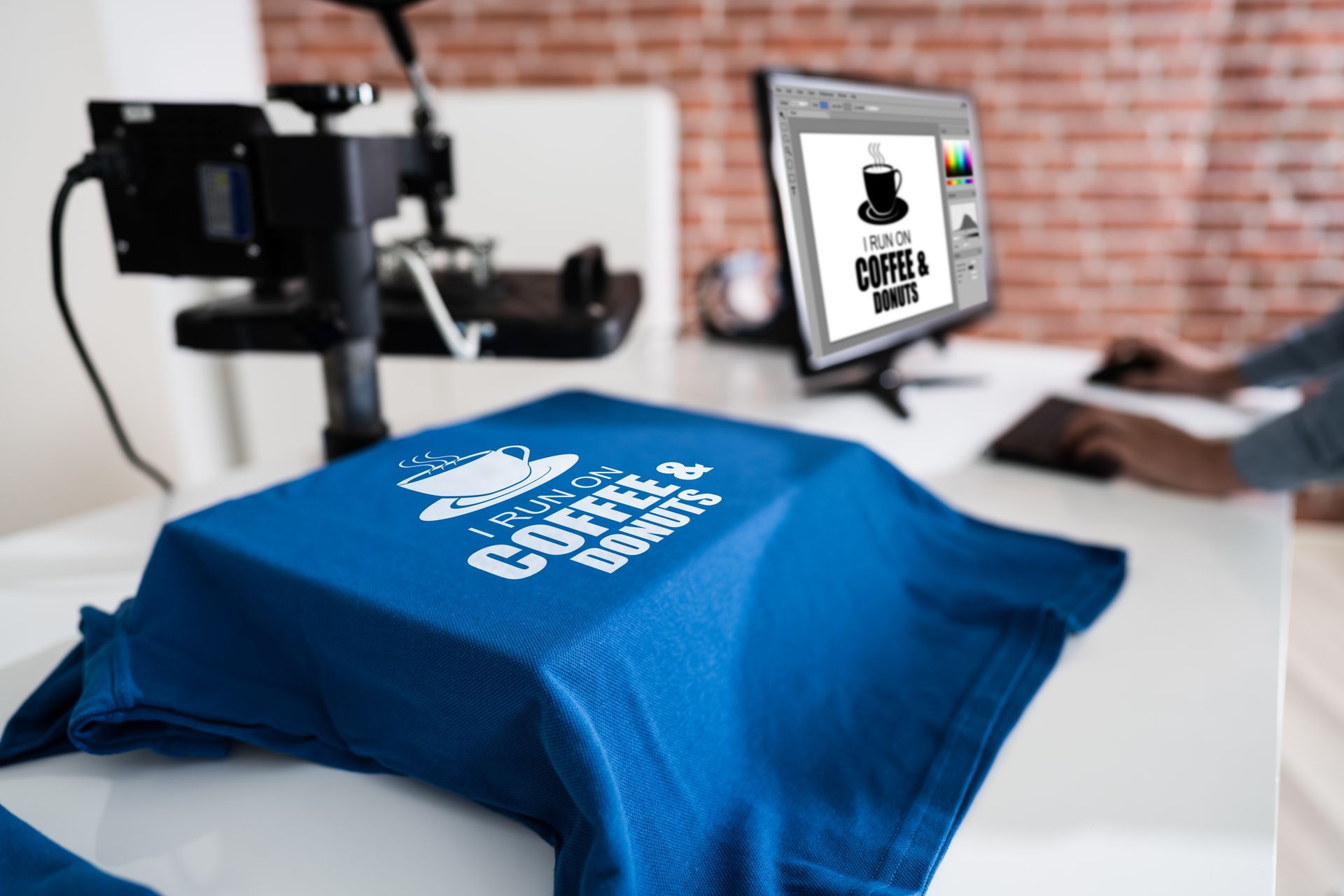 A blue t-shirt being customized with a coffee and donuts design using a heat press machine on a white desk.