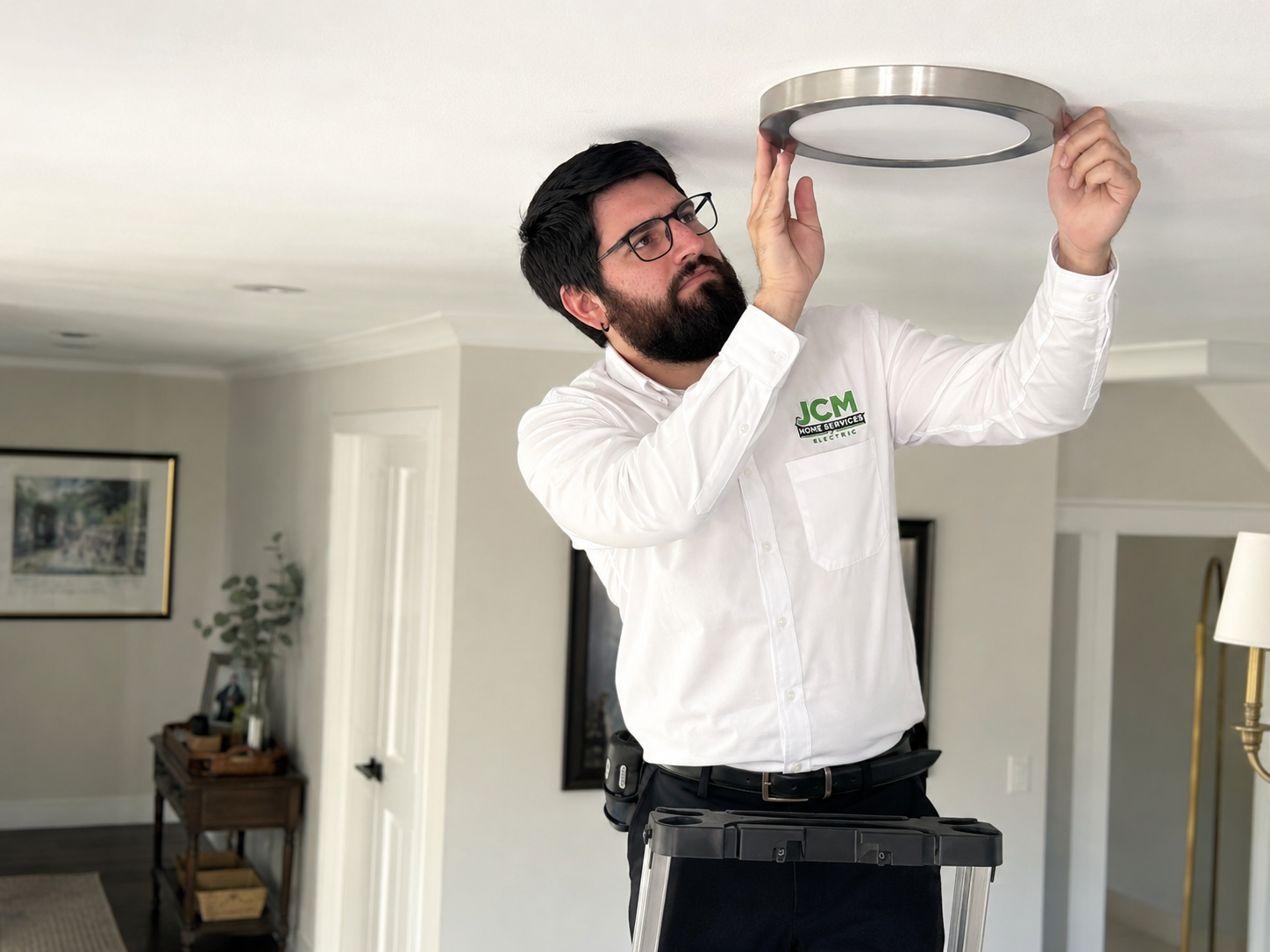 Man installing a circular ceiling light in a white room, standing on a ladder.